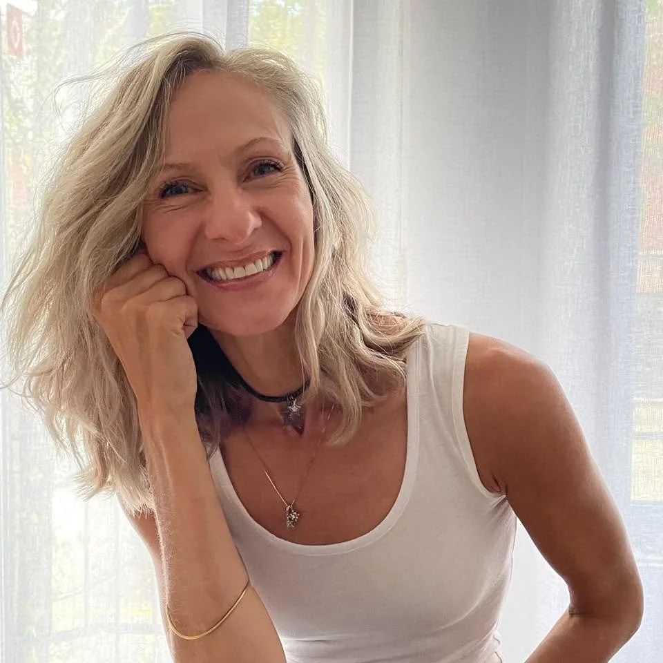 Une femme souriante aux cheveux blonds, portant un t-shirt blanc, avec des bijoux et des colliers, devant un rideau blanc.