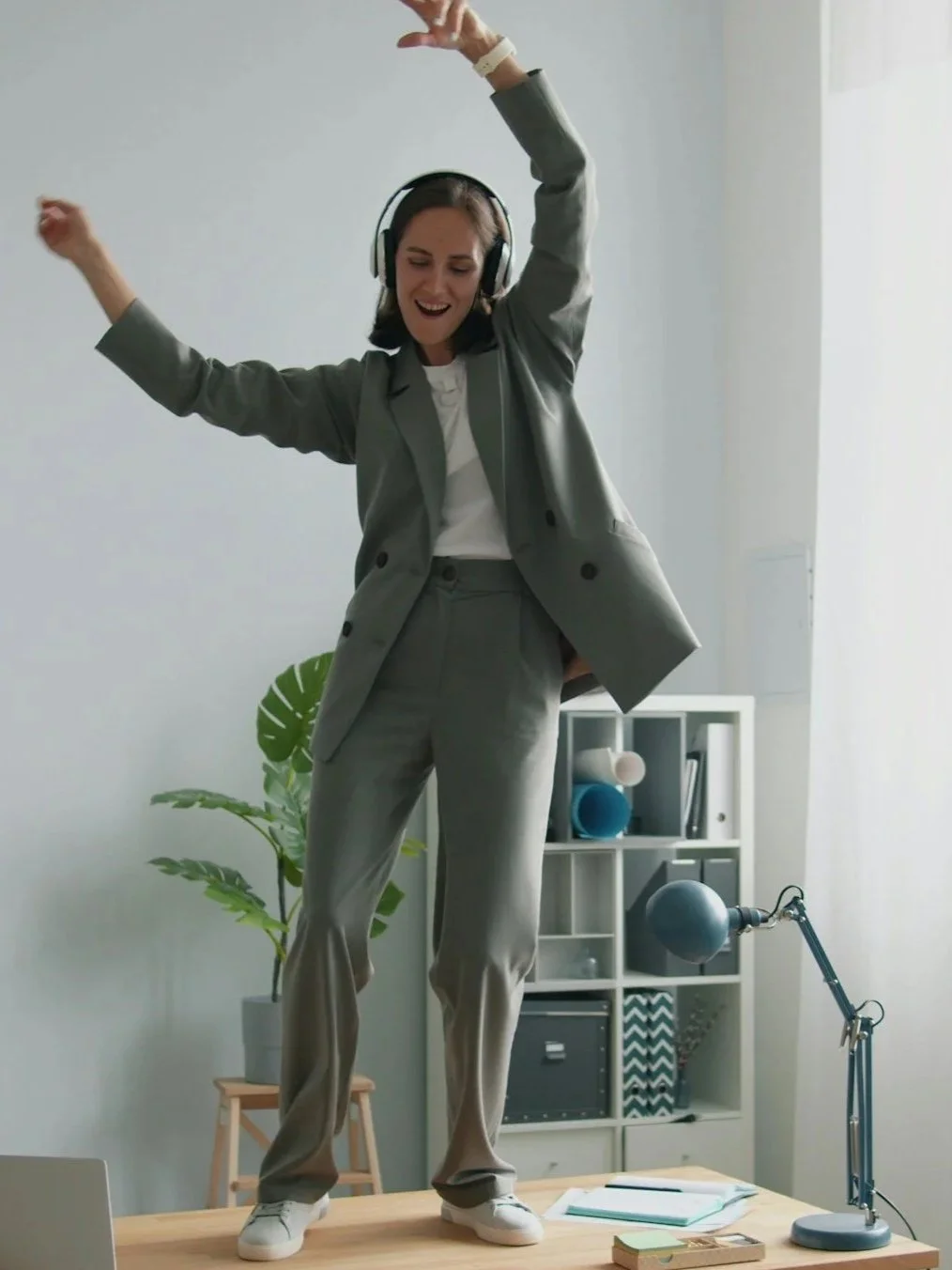 Femme souriante en costume gris, dansant sur un bureau en milieu de bureau moderne. Voir l'offre aux entreprises de Racines & Mouvements