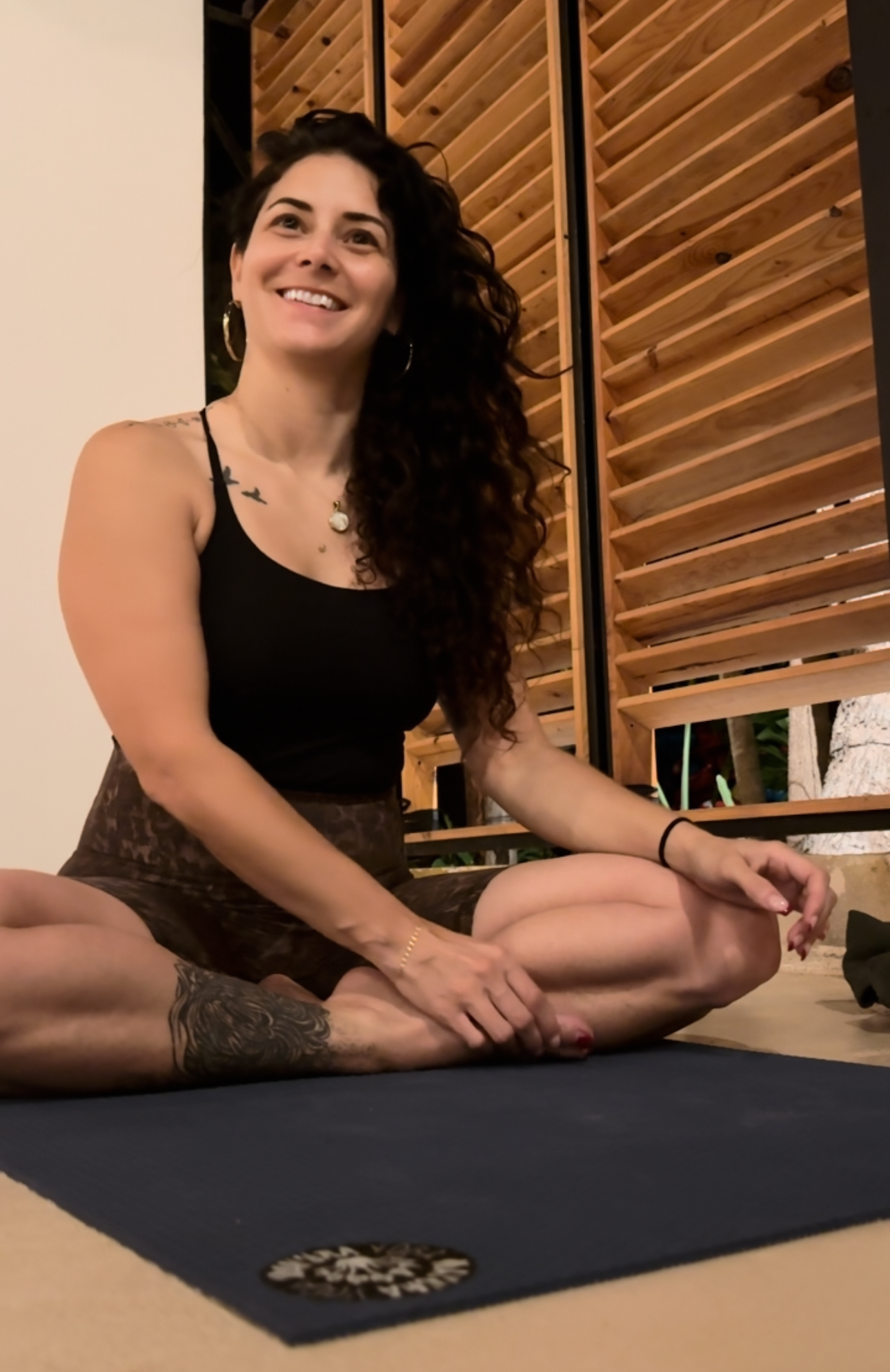 Femme souriante pratiquant le yoga en méditation, assise en position loto, sur un tapis noir, avec des murs en bois en arrière-plan.