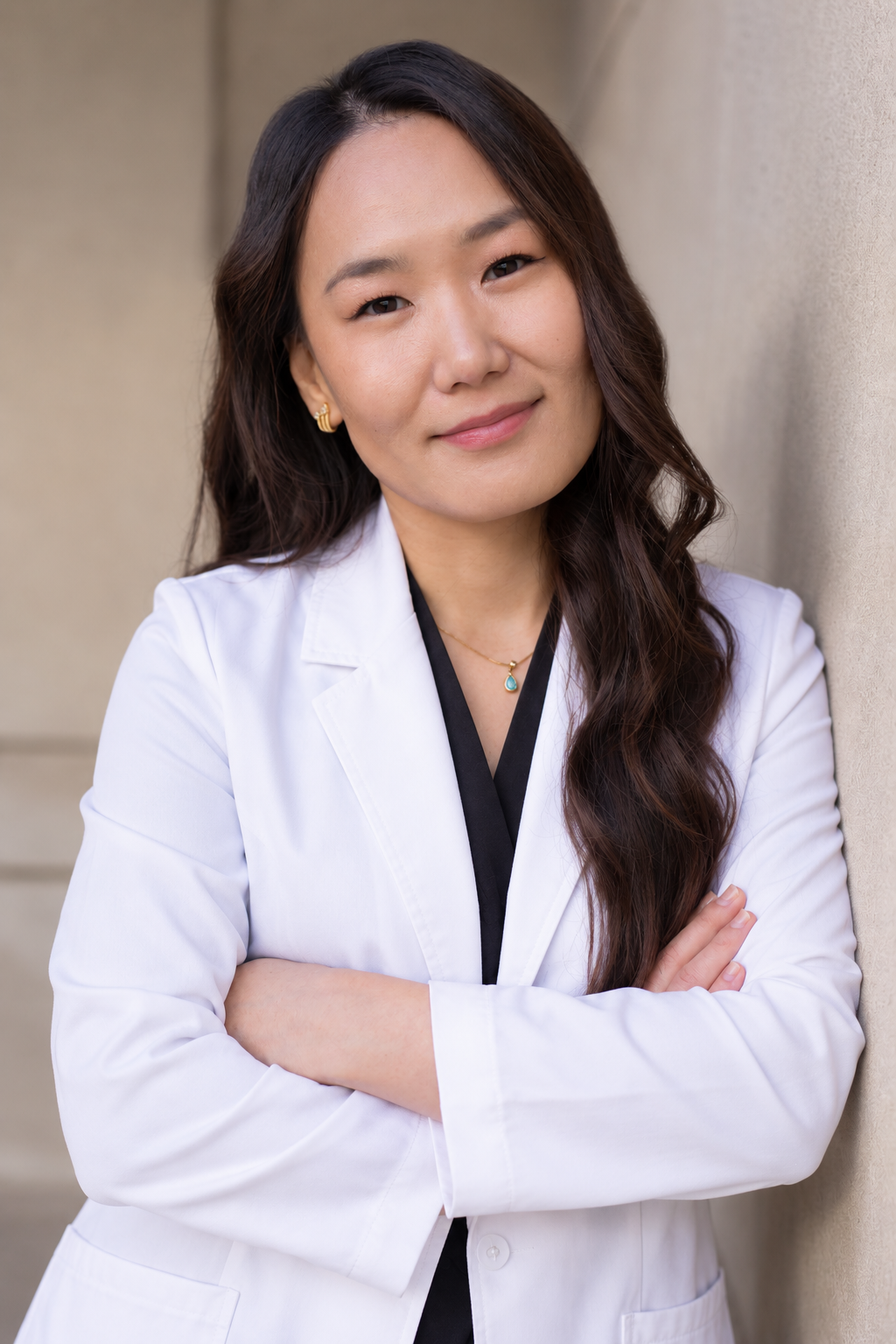 A woman with long wavy dark hair wearing a white lab coat and gold jewelry, standing against a beige wall, looking at the camera with a slight smile.