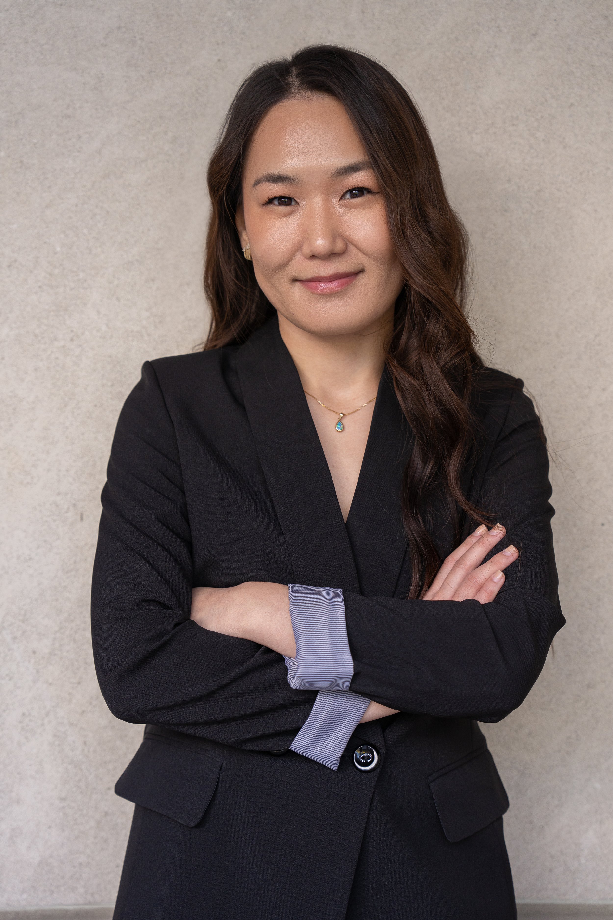Confident woman with long brown hair, wearing a black blazer and a necklace, standing with arms crossed against a light beige wall.
