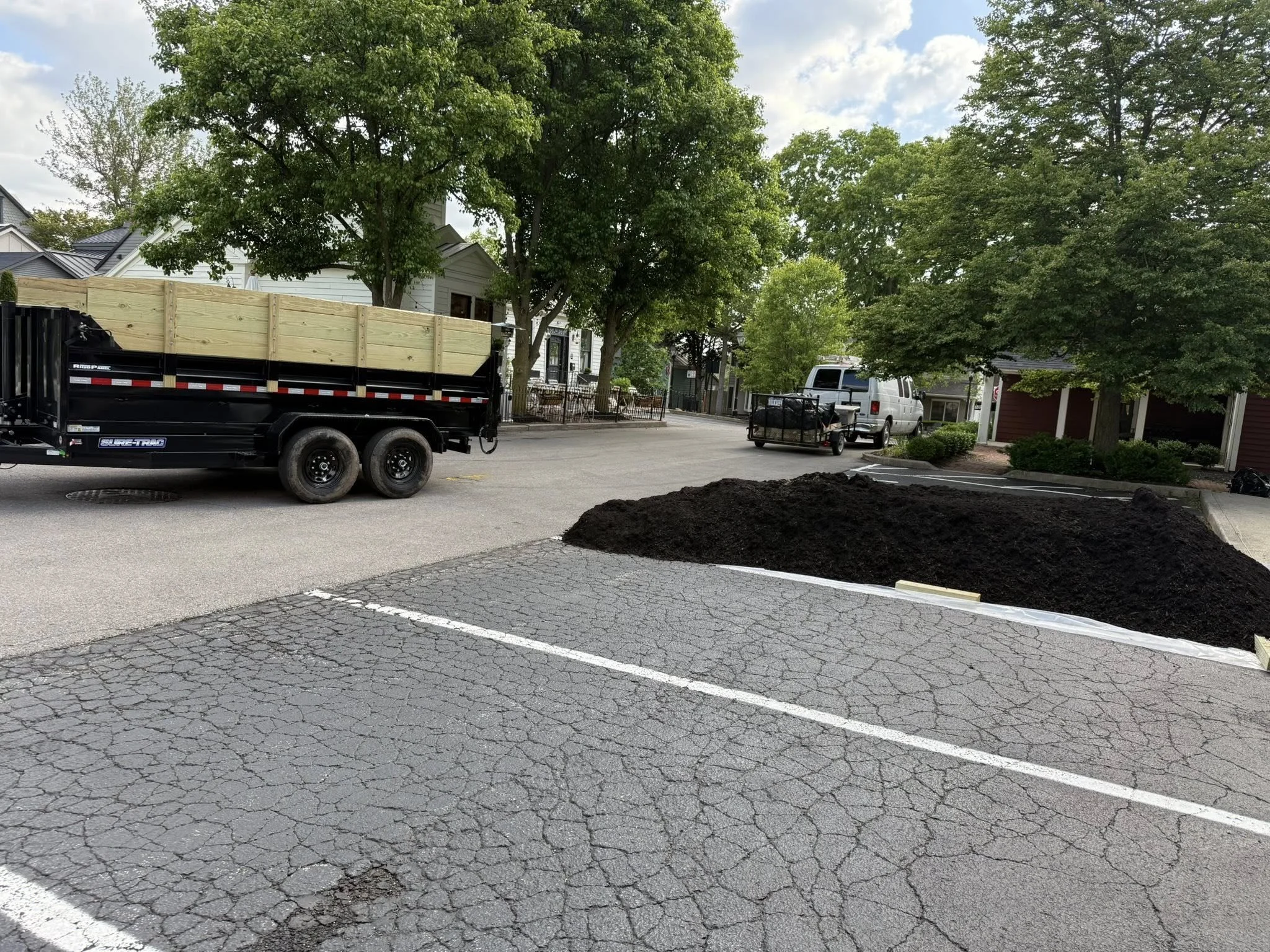 A parking lot with cracked pavement, a large pile of dirt, a black truck with a wooden fence load, trees, houses, parked vehicles, and a cloudy sky.