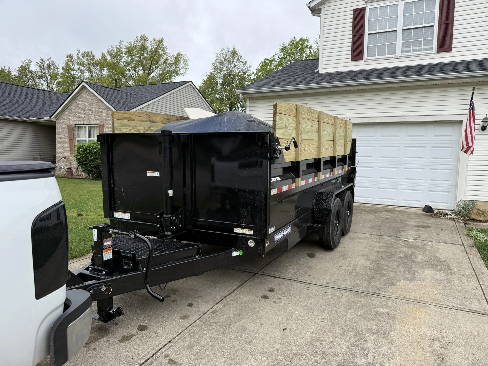 A black utility trailer parked in a driveway in front of a white garage with a closed door, attached to a white pickup truck. The trailer has wooden side panels, and a black dome-shaped cover on top. An American flag is mounted on the house to the right, with siding and a window with burgundy shutters. There are trees and neighboring houses in the background.