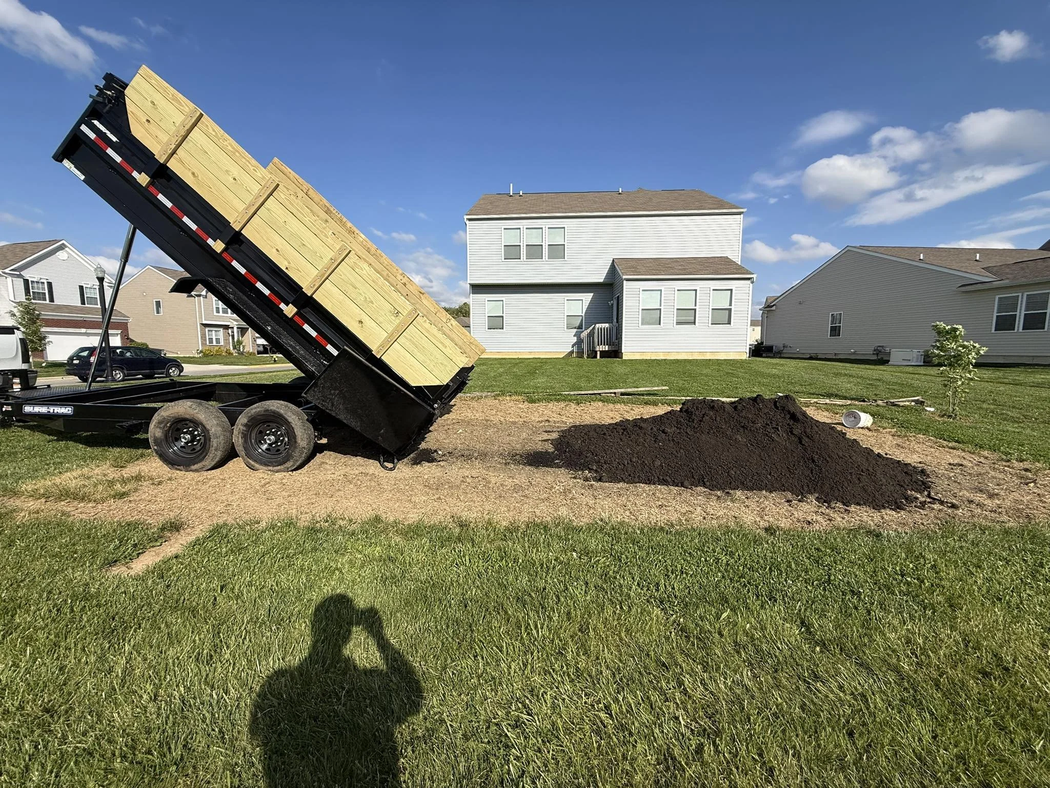 A trailer dumping a load of dark soil in a backyard with a house and neighboring homes in the background, under a blue sky with clouds.