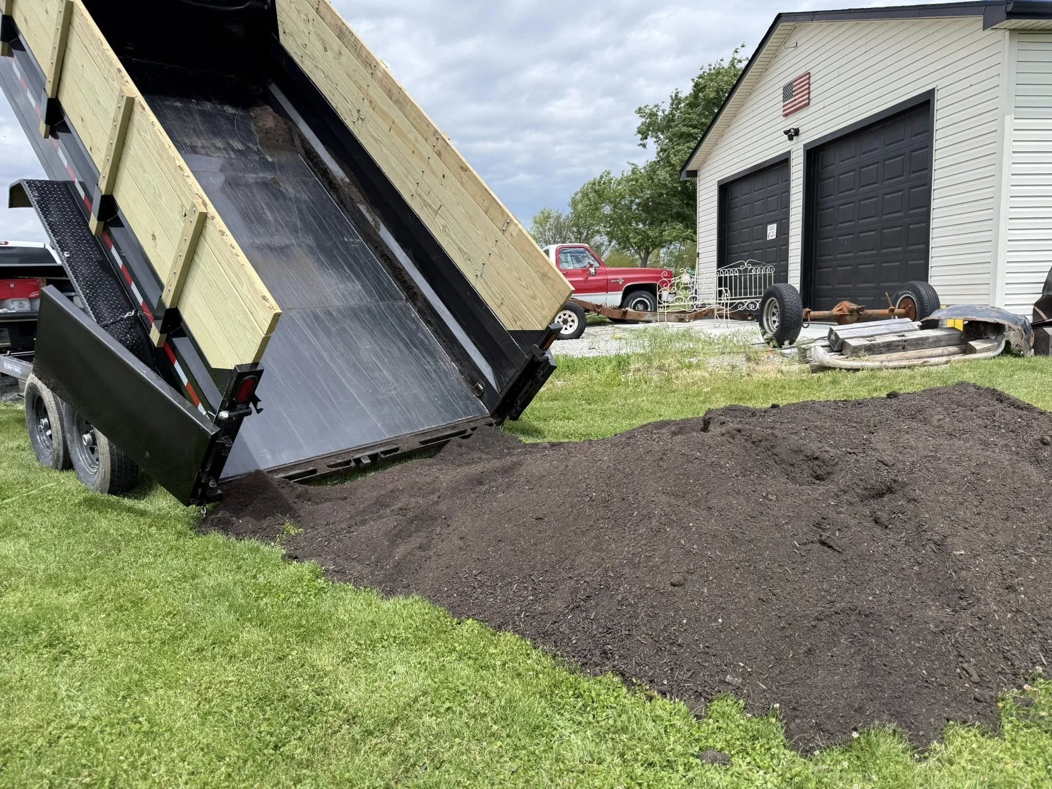A trailer dumping dark soil in the yard next to a white garage with an American flag decoration. A red pickup truck is visible in the background.