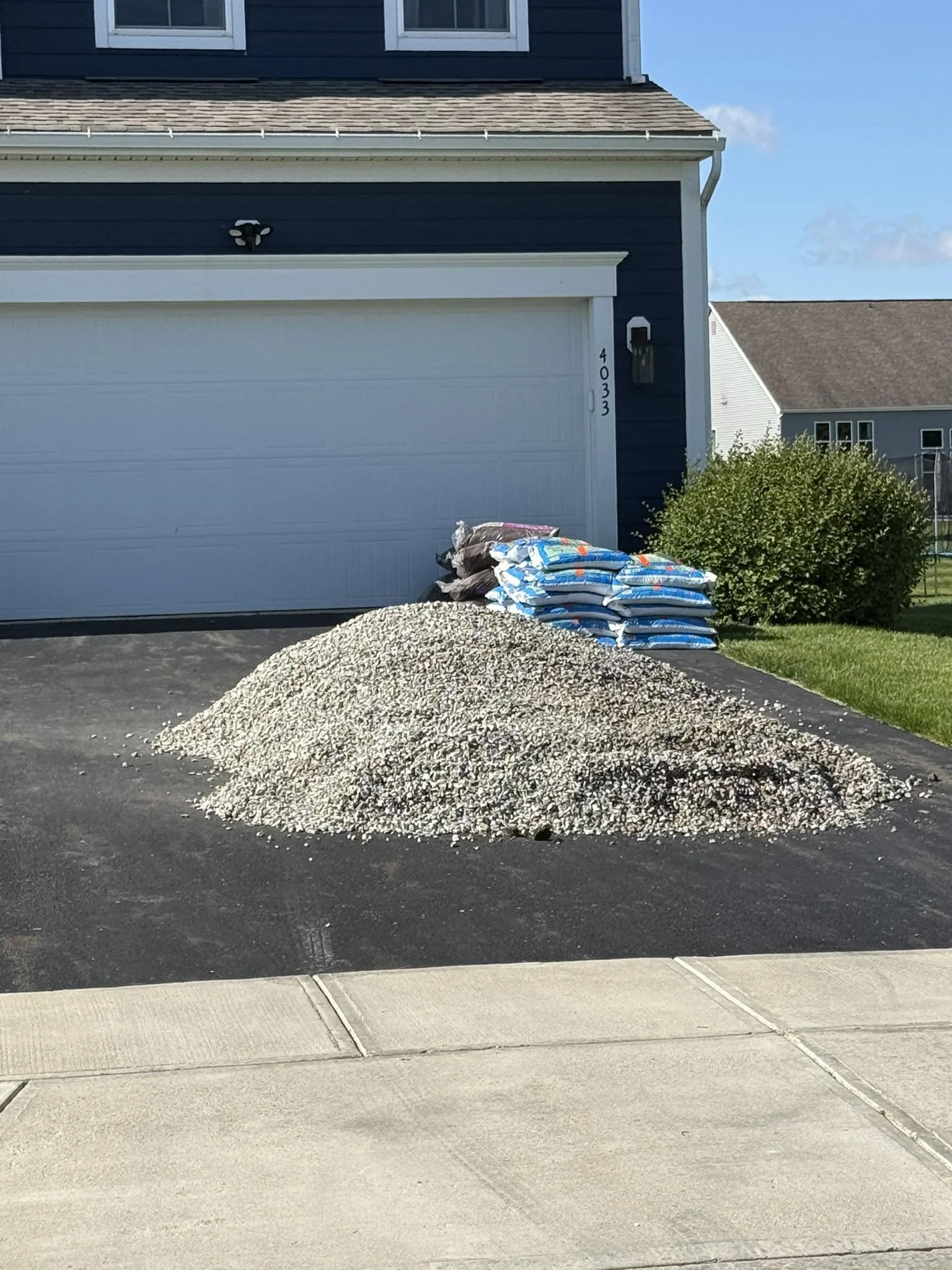 A pile of white and brown gravel and multiple bags of landscaping supplies in front of a blue house with white trim, next to a green shrub. The house has a garage with house number 4033 and an outdoor light fixture.