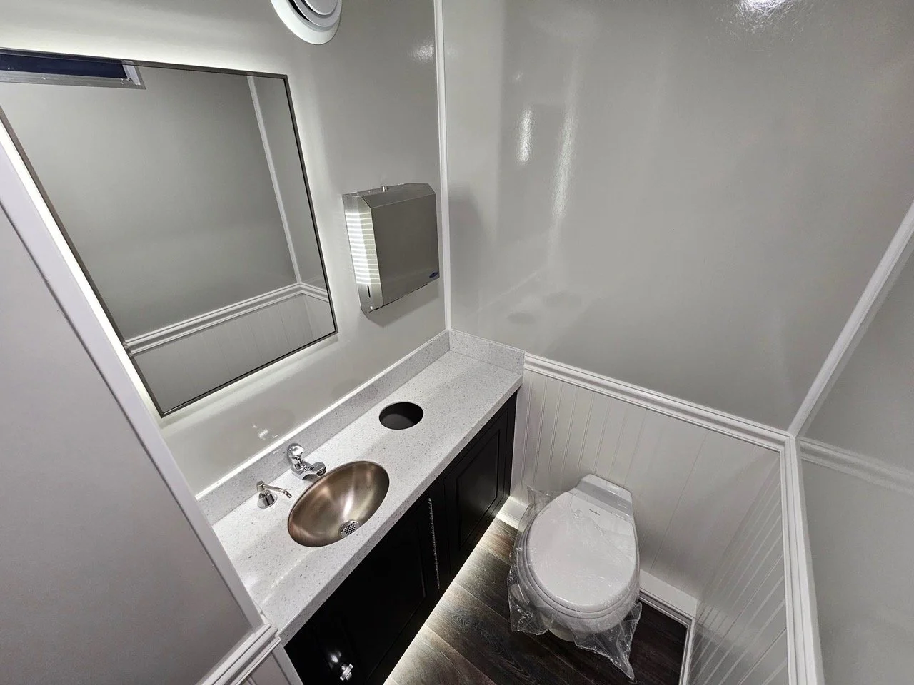 Small bathroom with a white countertop, a undermount sink, and a large mirror above it. A wall-mounted stainless steel paper towel dispenser is next to the mirror. The bathroom also has a toilet wrapped in plastic, dark wood flooring, and white beadboard wall paneling.