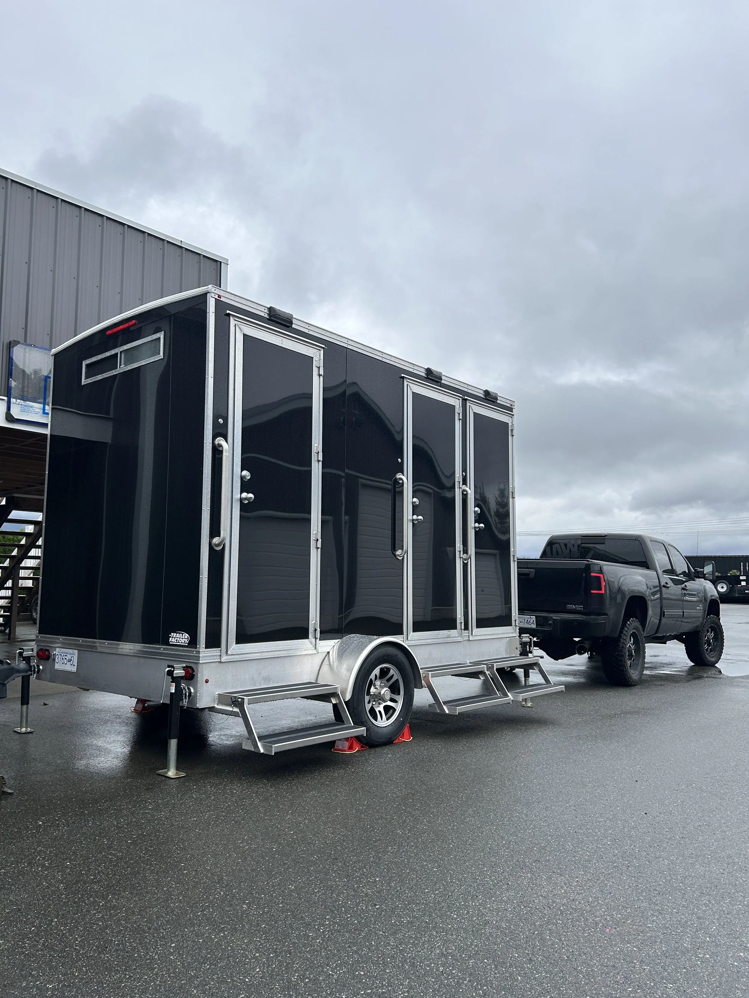 Black trailer attached to a black pickup truck parked on a wet asphalt lot under gray cloudy sky with a gray building in the background.