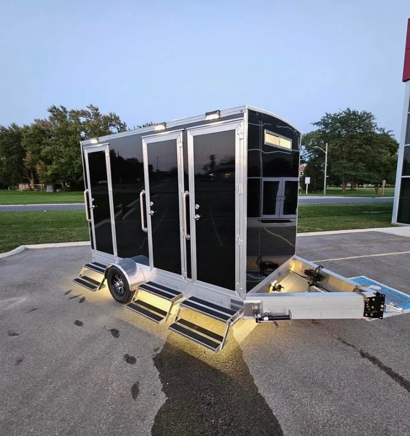 A black enclosed trailer with silver trim and steps on its side, parked on a paved lot at dusk with trees and a street in the background.