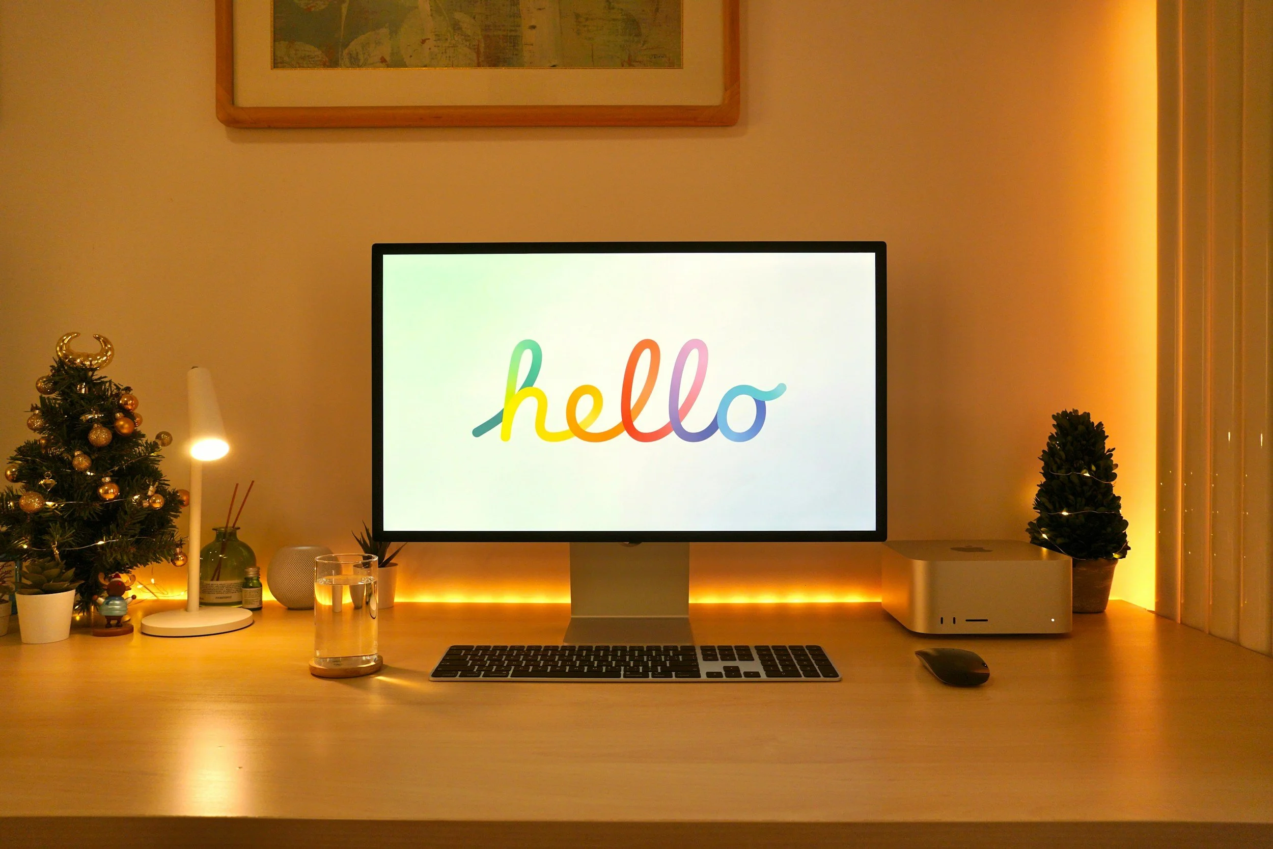A computer monitor displays the word 'hello' in colorful, cursive letters. The setup includes a keyboard, mouse, a glass of water, and decorative items like a small Christmas tree with ornaments, candles, and plants, all lit with warm ambient lighting.