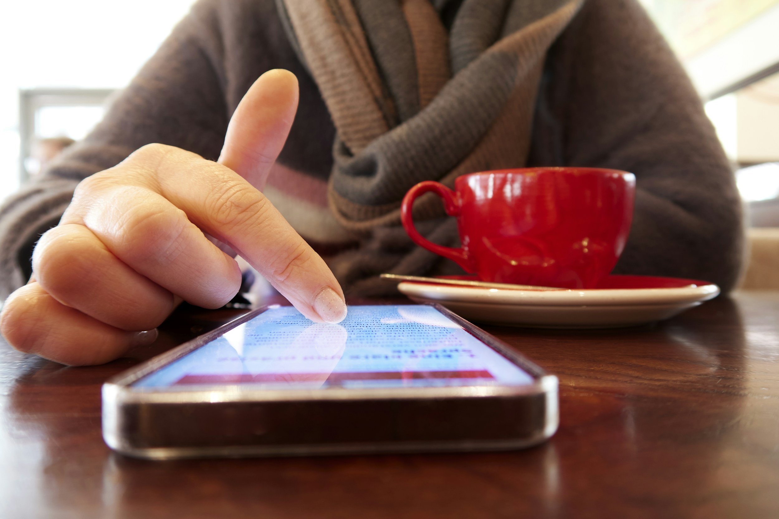 A person is using a smartphone at a table with a red coffee cup and saucer. The focus is on their hand and phone, with the coffee cup in the background.