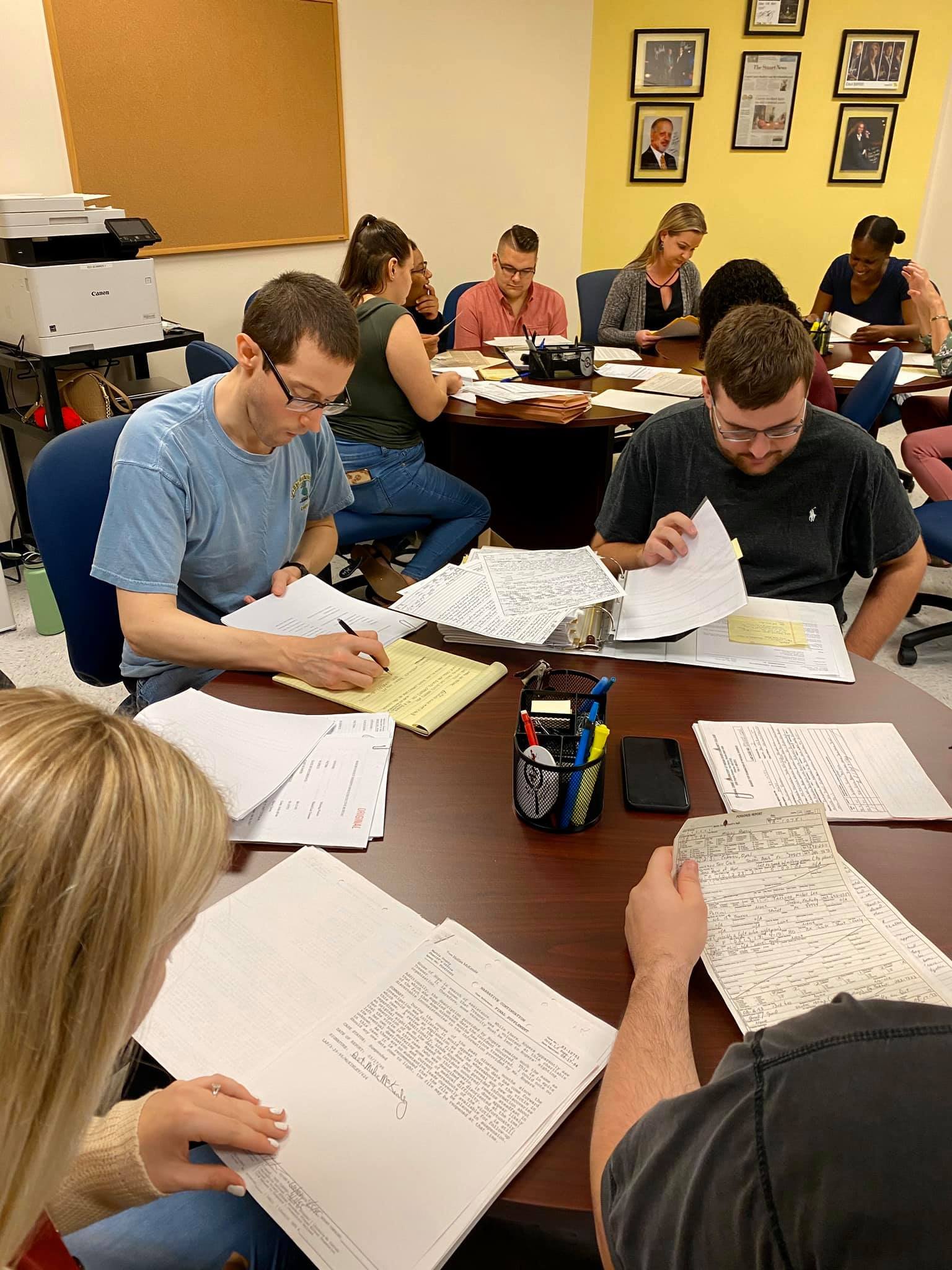 A group of people sitting around a table, reviewing documents and paperwork, in a meeting room with framed photos and articles on the yellow wall.