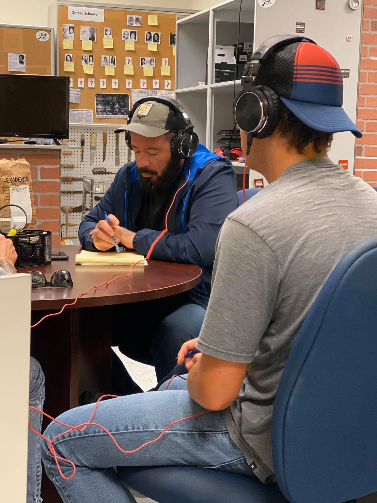 Two men sitting at a table, wearing headphones, with a corkboard with photos and notes, and tools on the wall behind them.