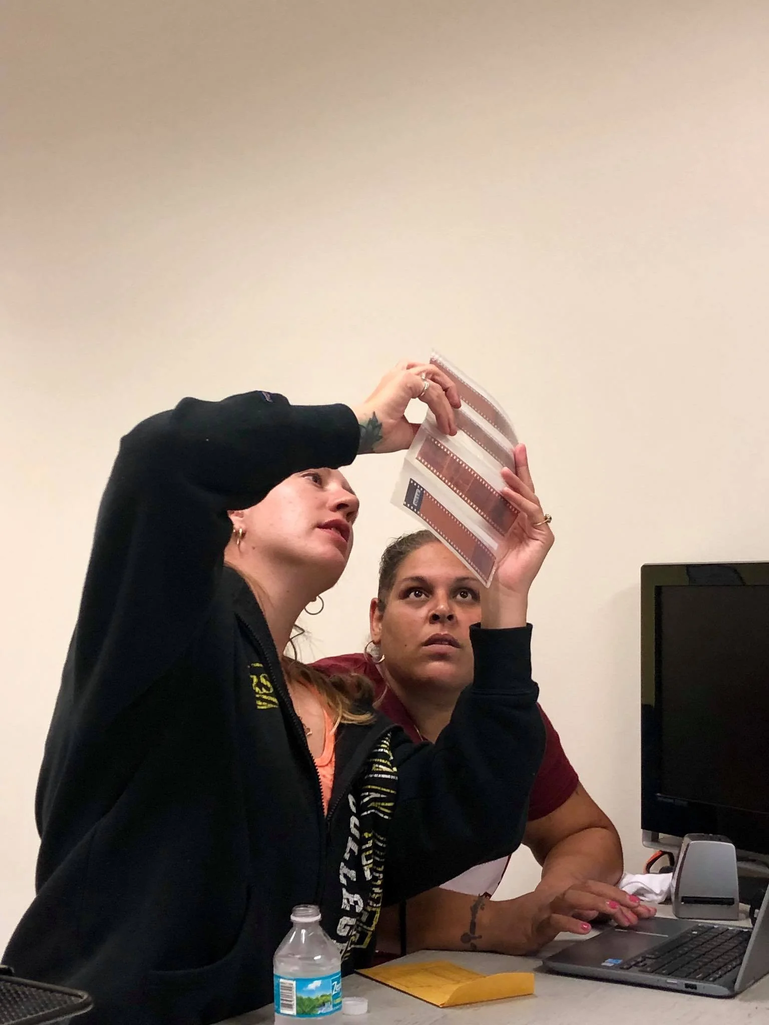 Two women are working together on a computer. One woman is standing and holding a film strip, while the other is sitting and typing on the laptop. On the table, there is a water bottle, a yellow envelope, and some papers.