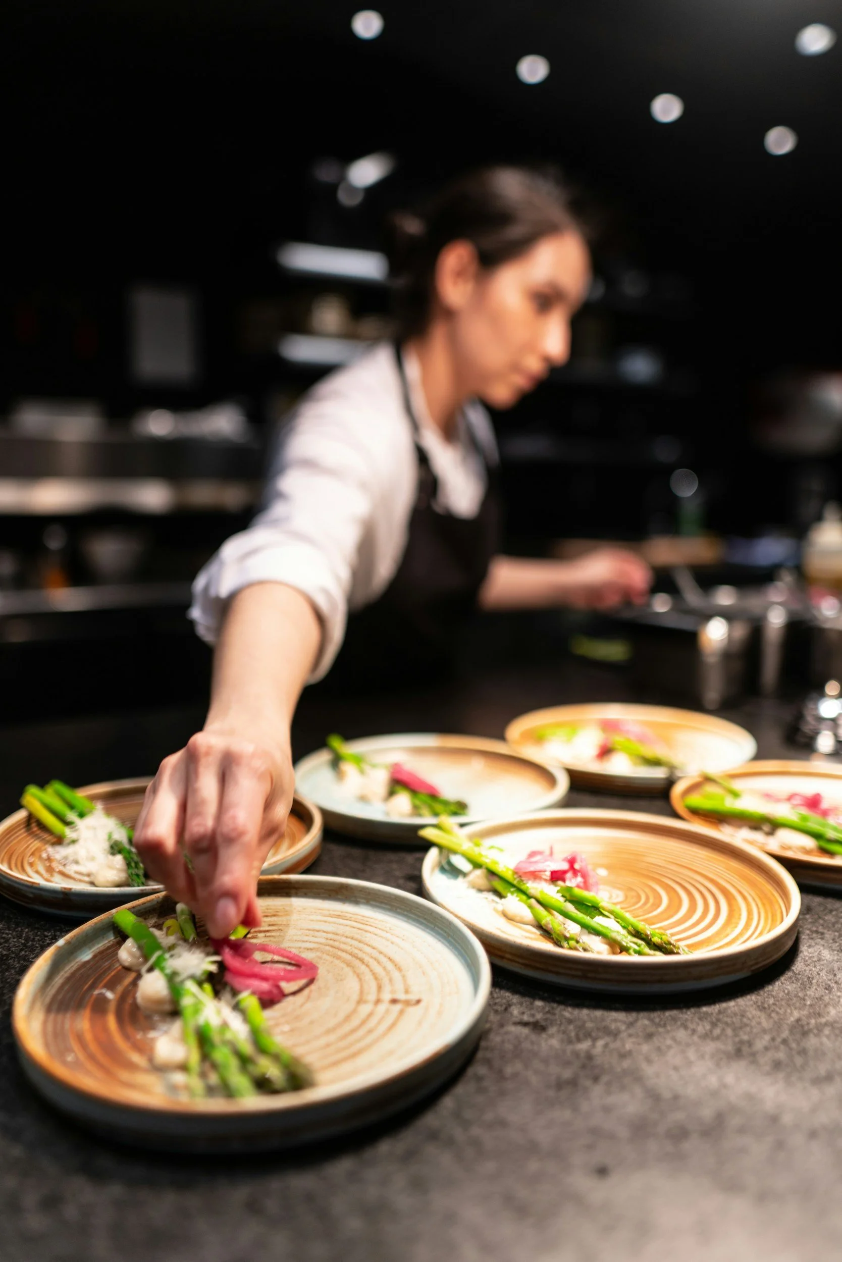 A chef arranging dishes with asparagus and thin slices of meat on decorative ceramic plates in a professional kitchen.