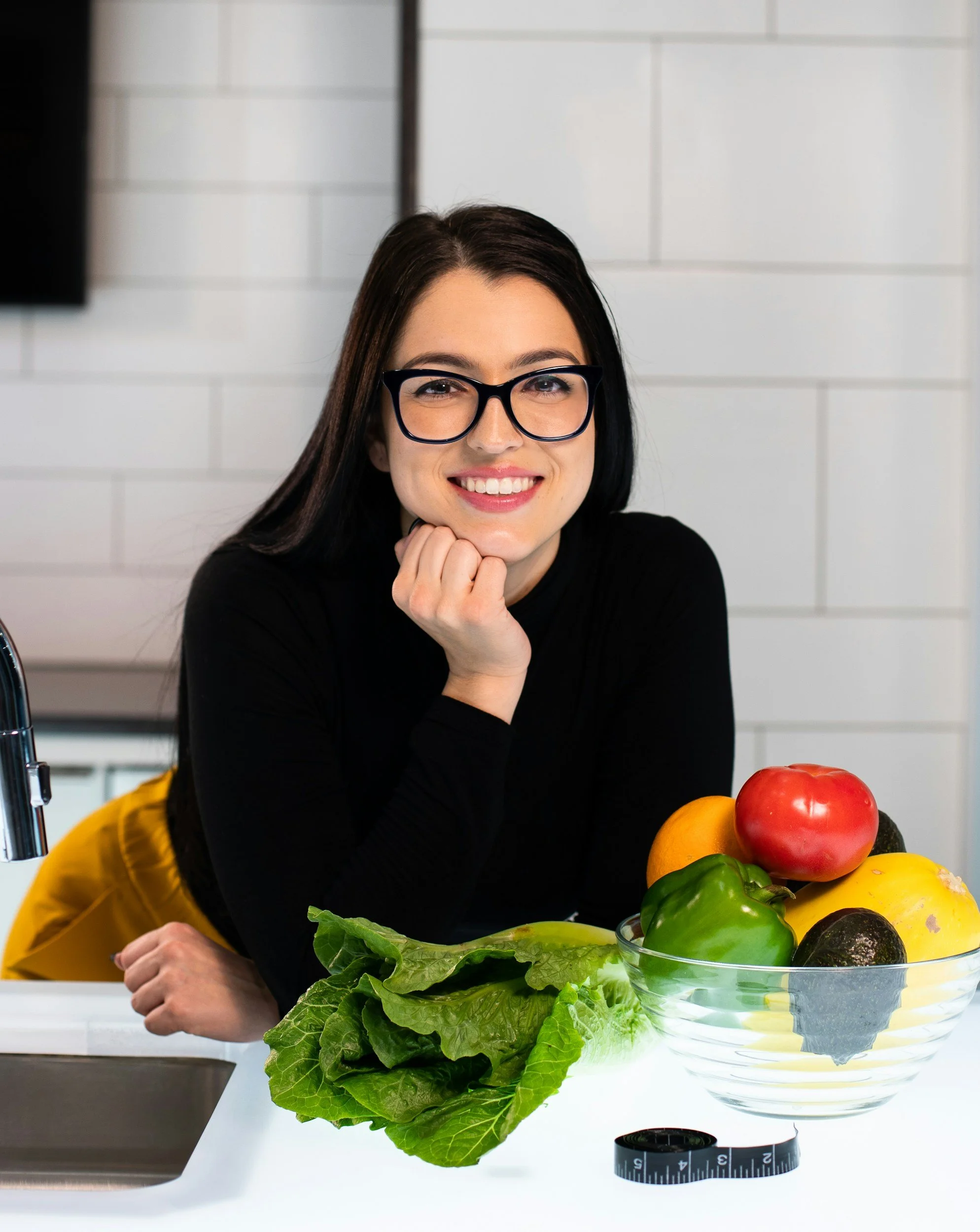 A woman with dark hair, glasses, and a black top sitting at a table with a large glass bowl of colorful bell peppers and lettuce in front of her in a kitchen with white tiled walls.