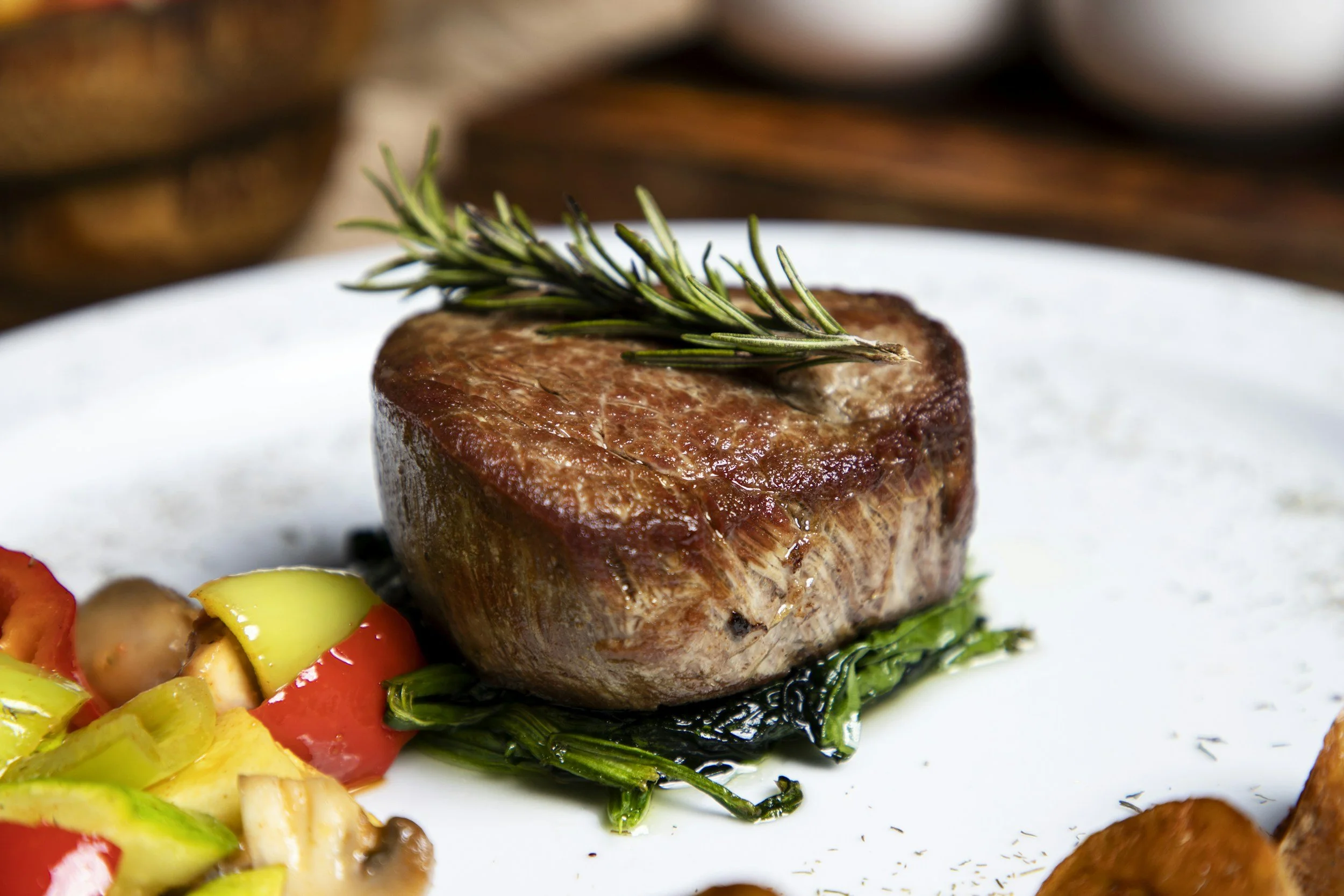 A medium cooked steak topped with a sprig of rosemary, served with chopped vegetables including yellow and red bell peppers and mushrooms.