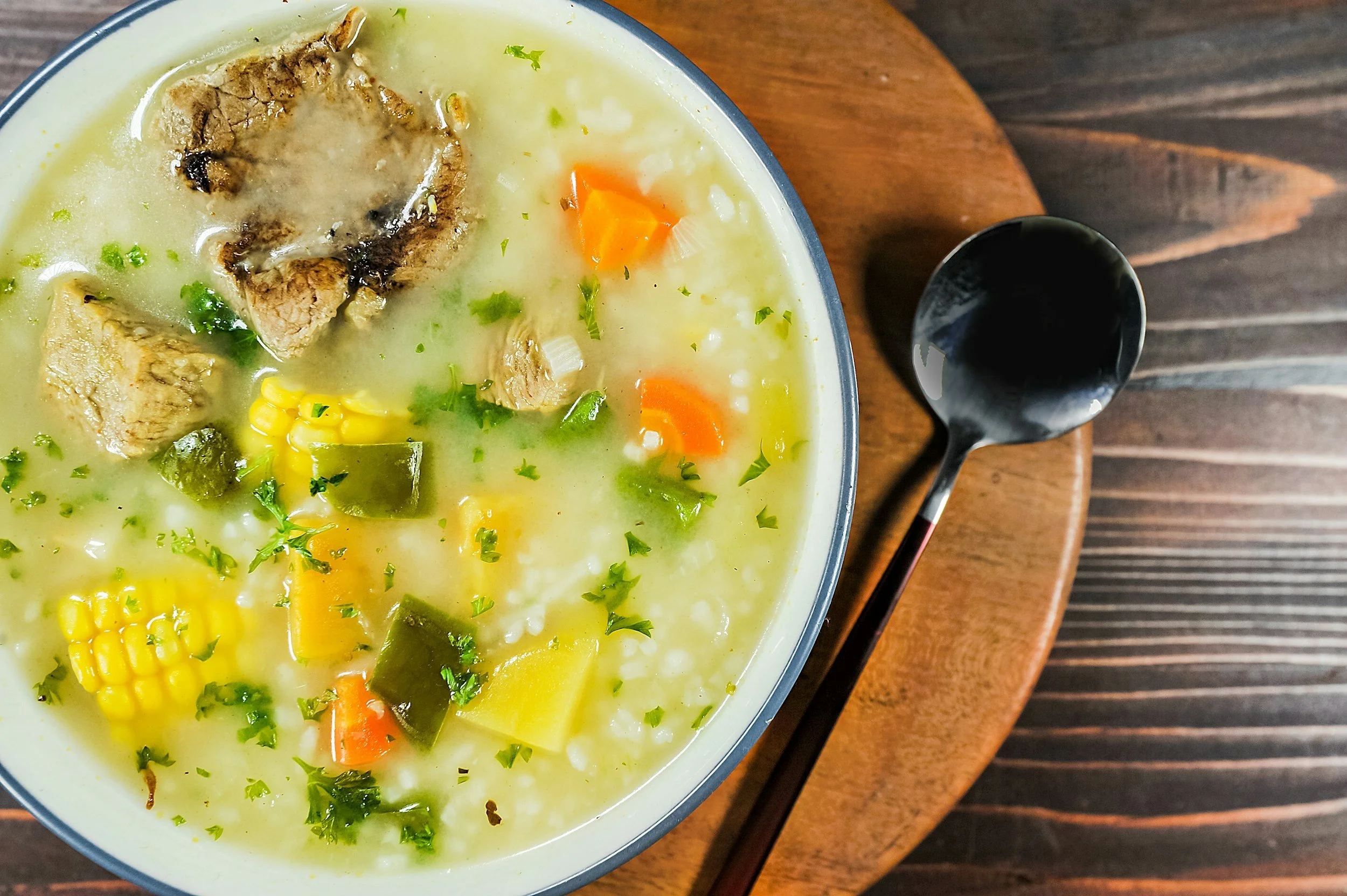 A bowl of creamy chicken and vegetable soup with corn, carrots, celery, and herbs on a wooden table with a black spoon beside it.