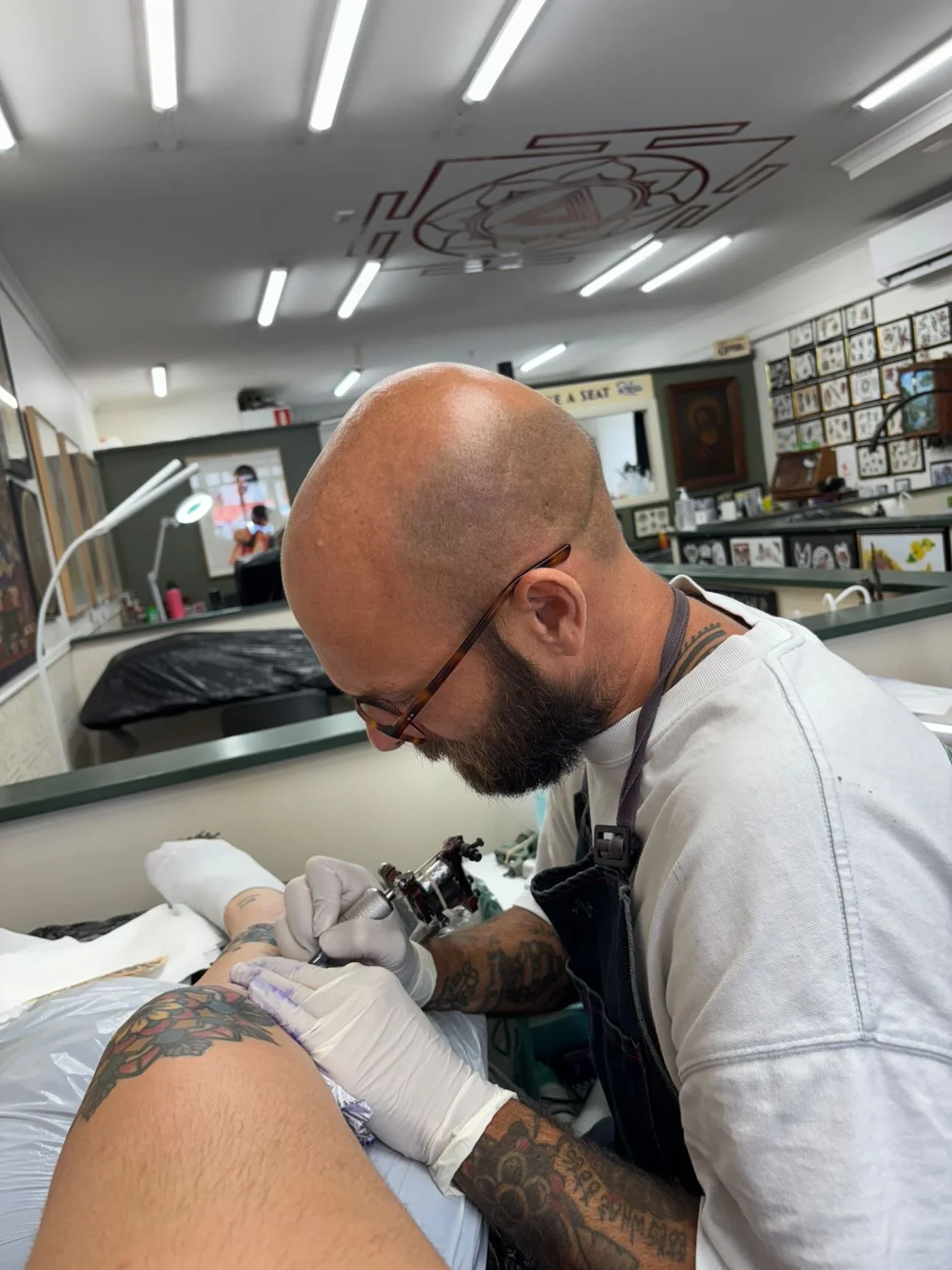 Tattoo artist working on a client's arm in a tattoo studio, with tattoo designs on the wall in the background.