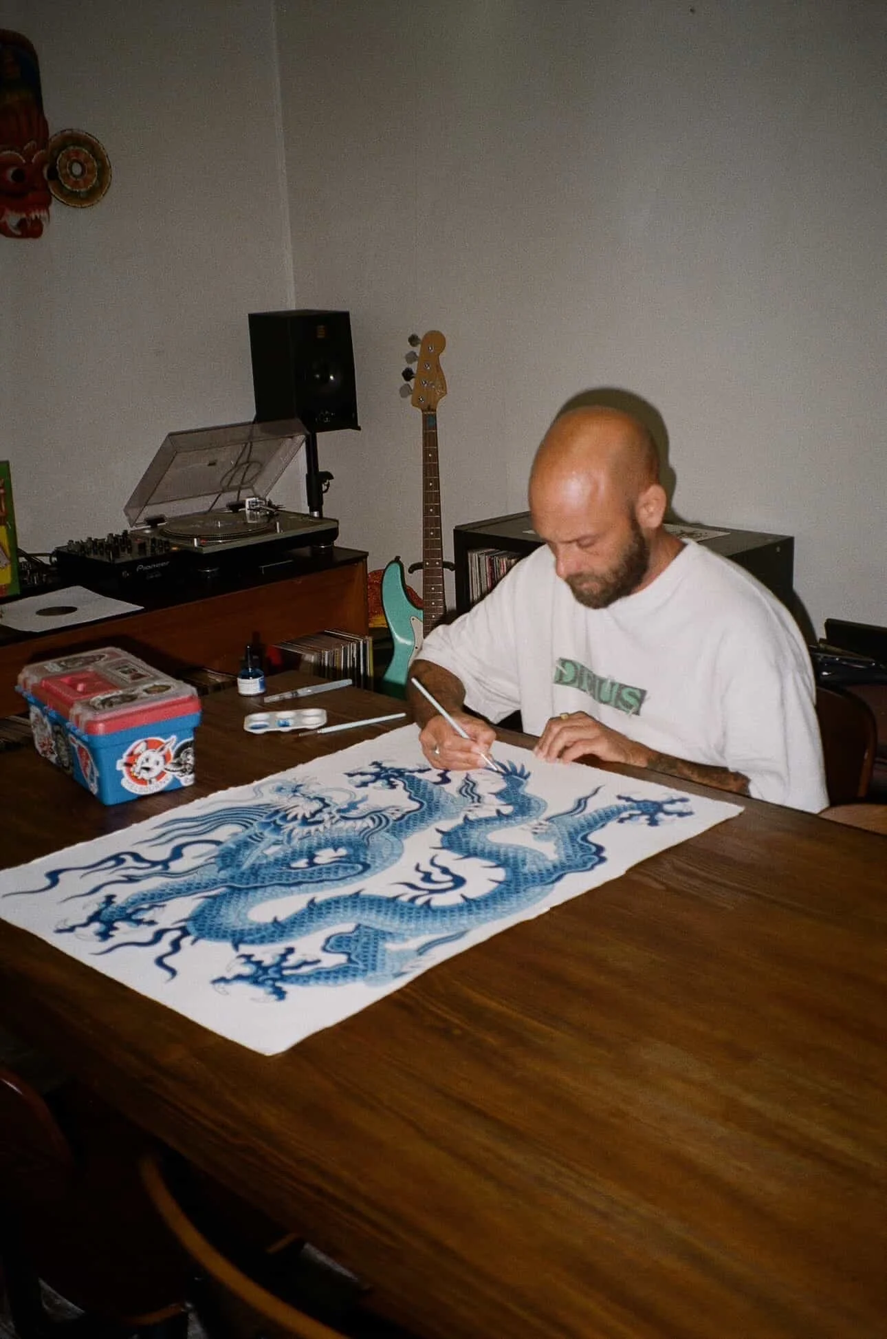 Man with a beard and bald head working on a detailed blue dragon drawing on white paper at a wooden table in a room with musical instruments, including a guitar and a turntable.