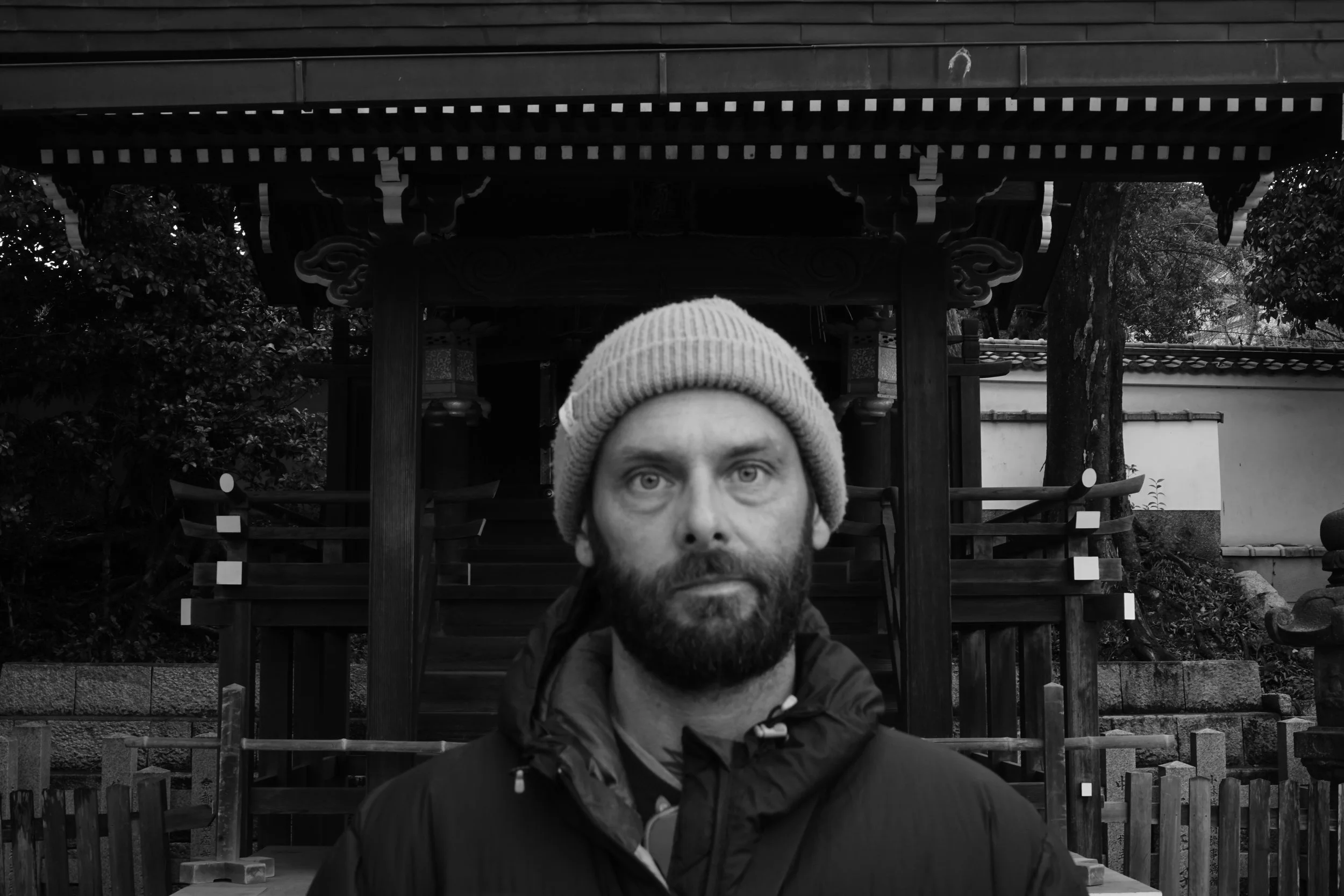 A man with a beard and a knit cap standing outdoors in front of a traditional Japanese temple gate, with trees and a wall in the background.