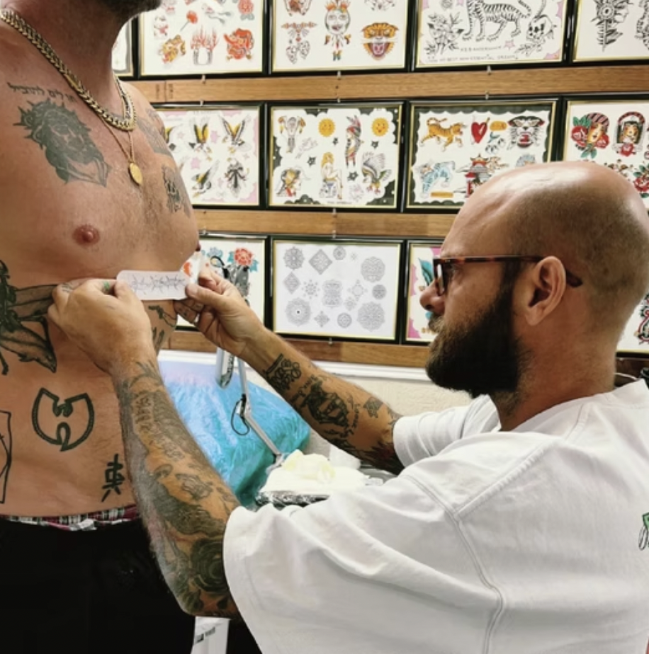 Tattoo artist applying a tattoo to a person's chest in a tattoo parlor with tattoo designs on the wall in the background.