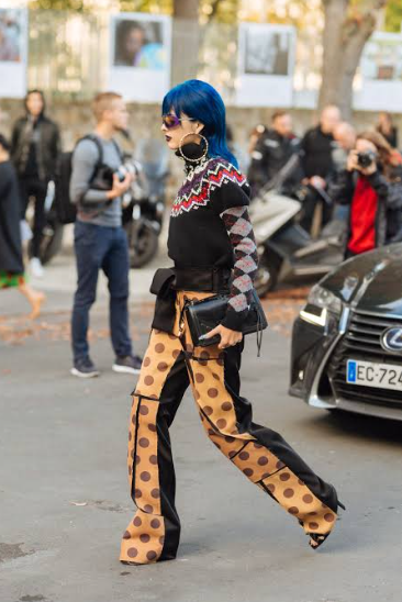 Person with vibrant blue hair, patterned sweater, and polka dot pants walking outdoors among other people and parked cars.