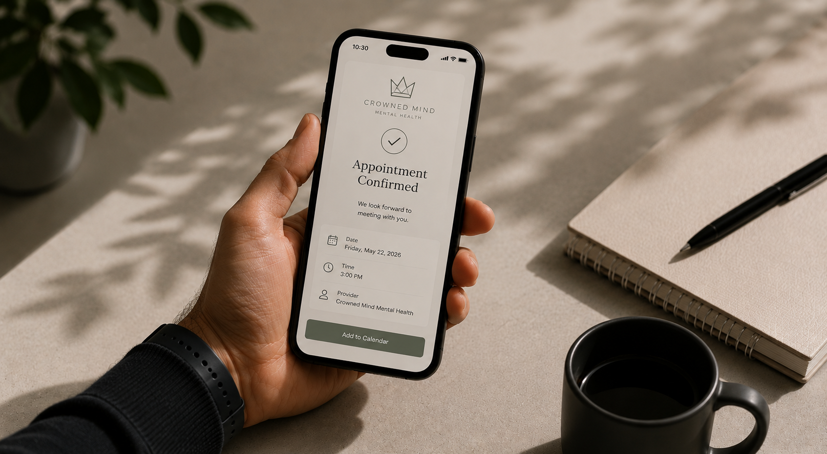 A person holding a smartphone displaying a mental health appointment confirmation screen. The screen shows the date, time, provider, and an option to add to the calendar. The person is wearing a black watch and is sitting at a desk with a notebook, a pen, a black mug, and a potted plant nearby.