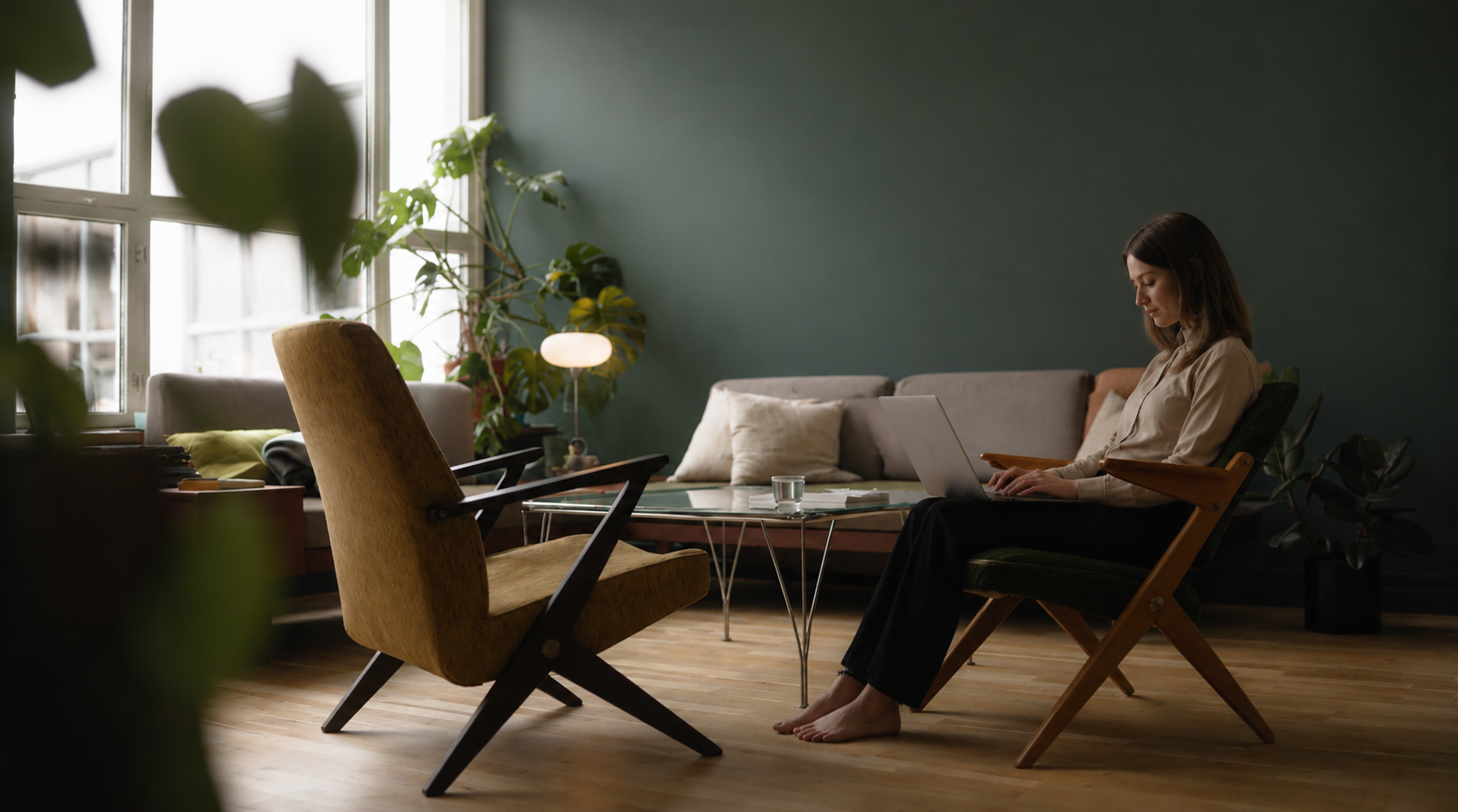 A woman working on a laptop in a cozy, well-lit living room with plants, a sofa, and wooden flooring.
