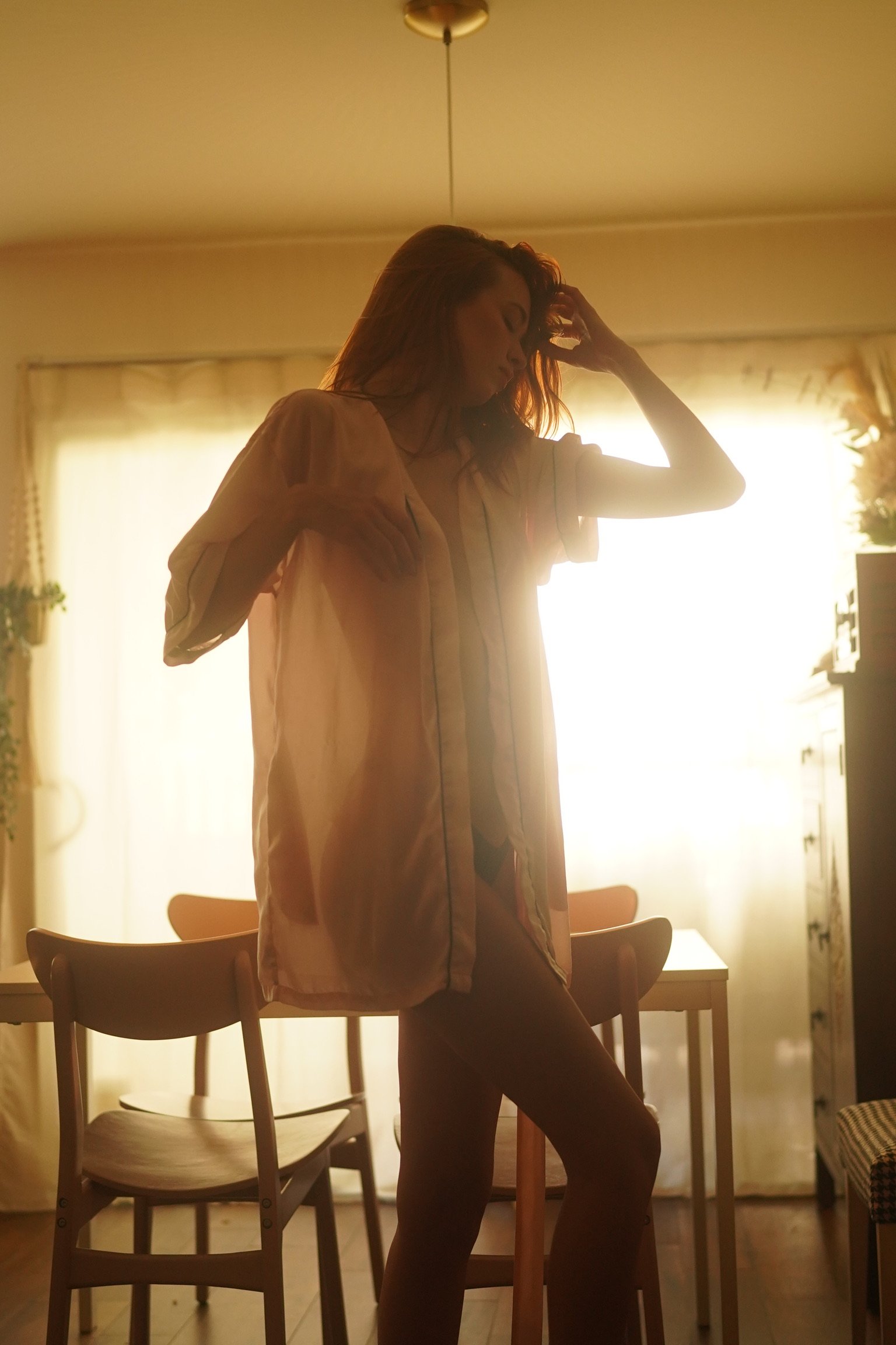 A woman with red hair standing in a sunlit room, touching her hair, wearing a loose shirt, with a dining table and chairs behind her.