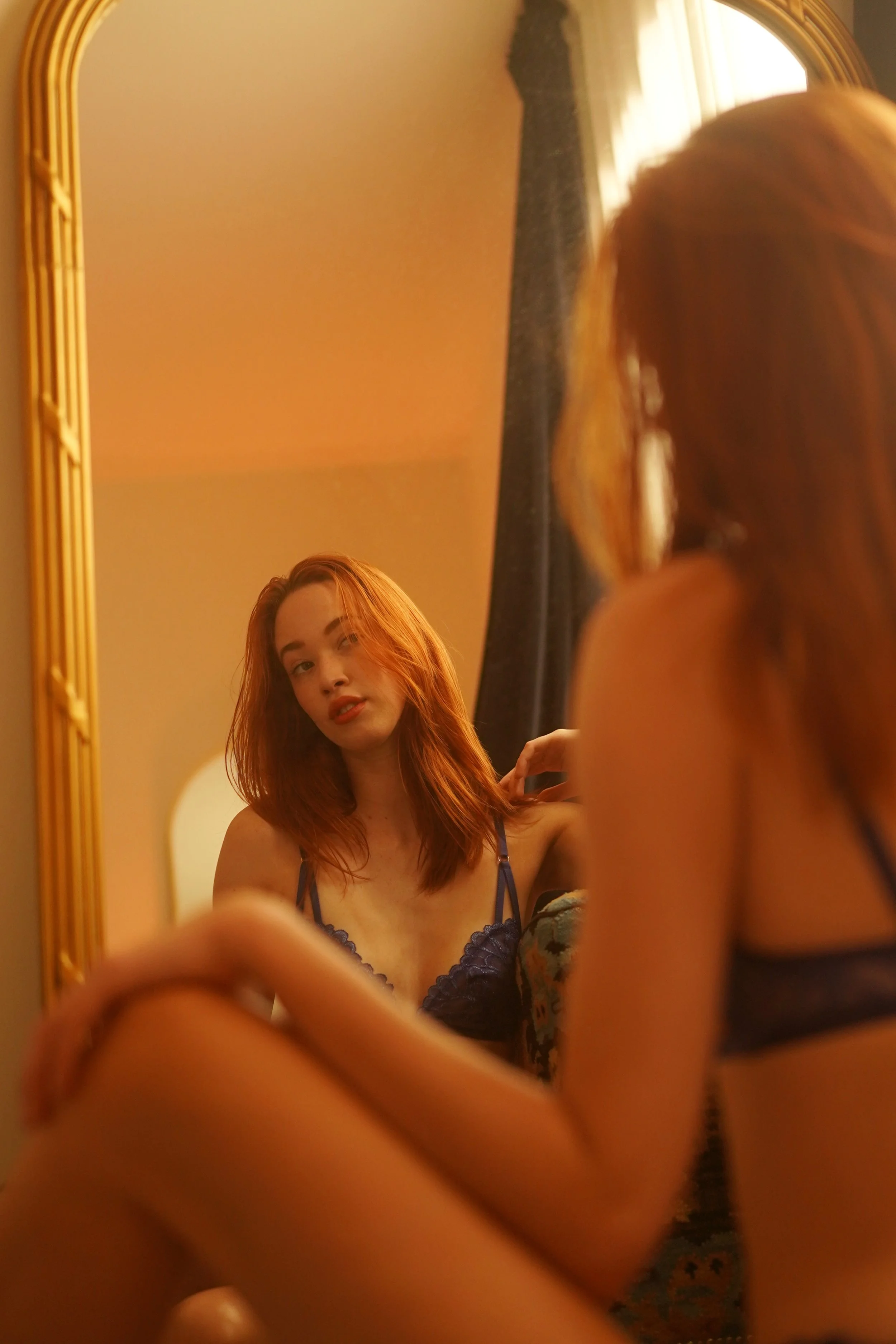 A young woman with red hair taking a selfie in a mirror, wearing a dark lace camisole, in a room with beige walls and a window with natural light.