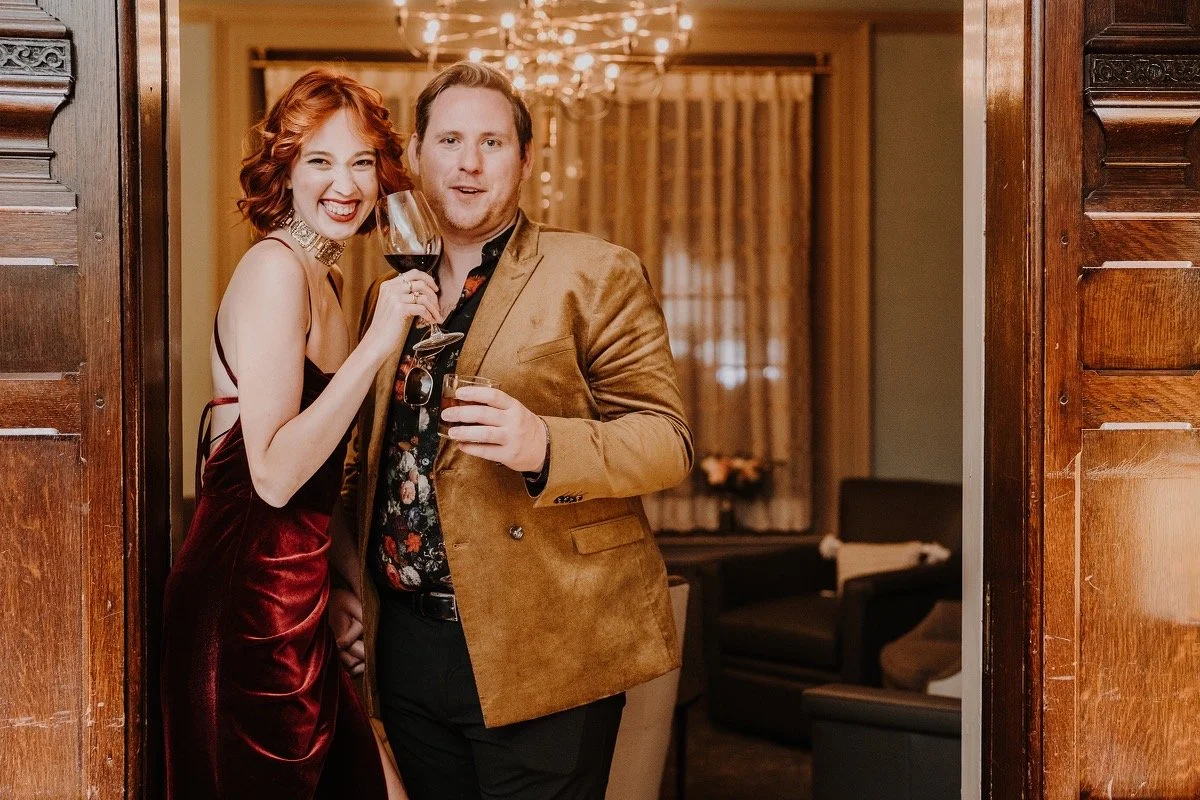 Hadley and Joe together, smiling and holding glasses of red wine, standing in a doorway of a warmly lit room with curtains and chandelier.