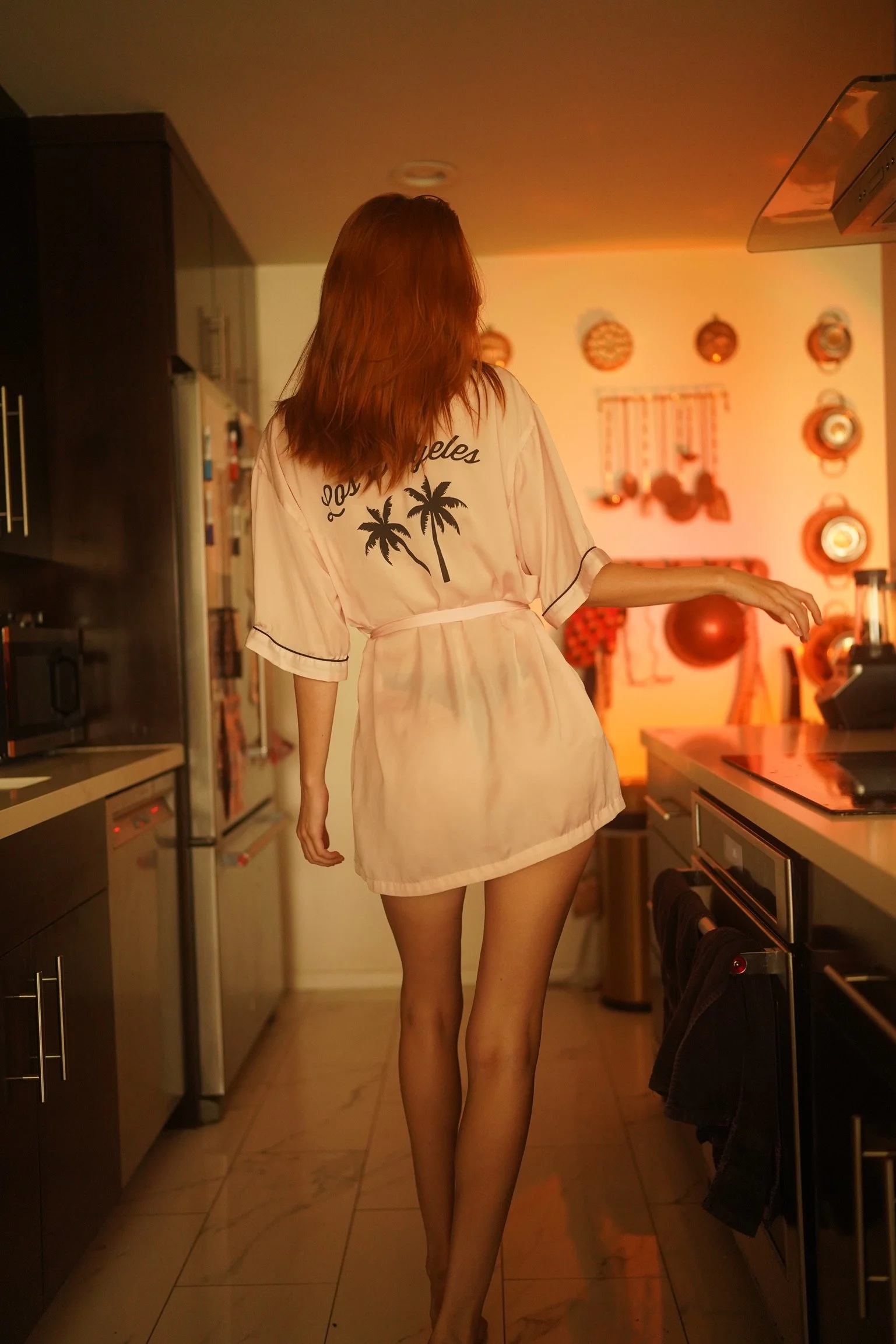 A woman with red hair wearing a light-colored robe with palm trees and "Los Angeles" printed on the back, standing in a kitchen with warm, dim lighting.