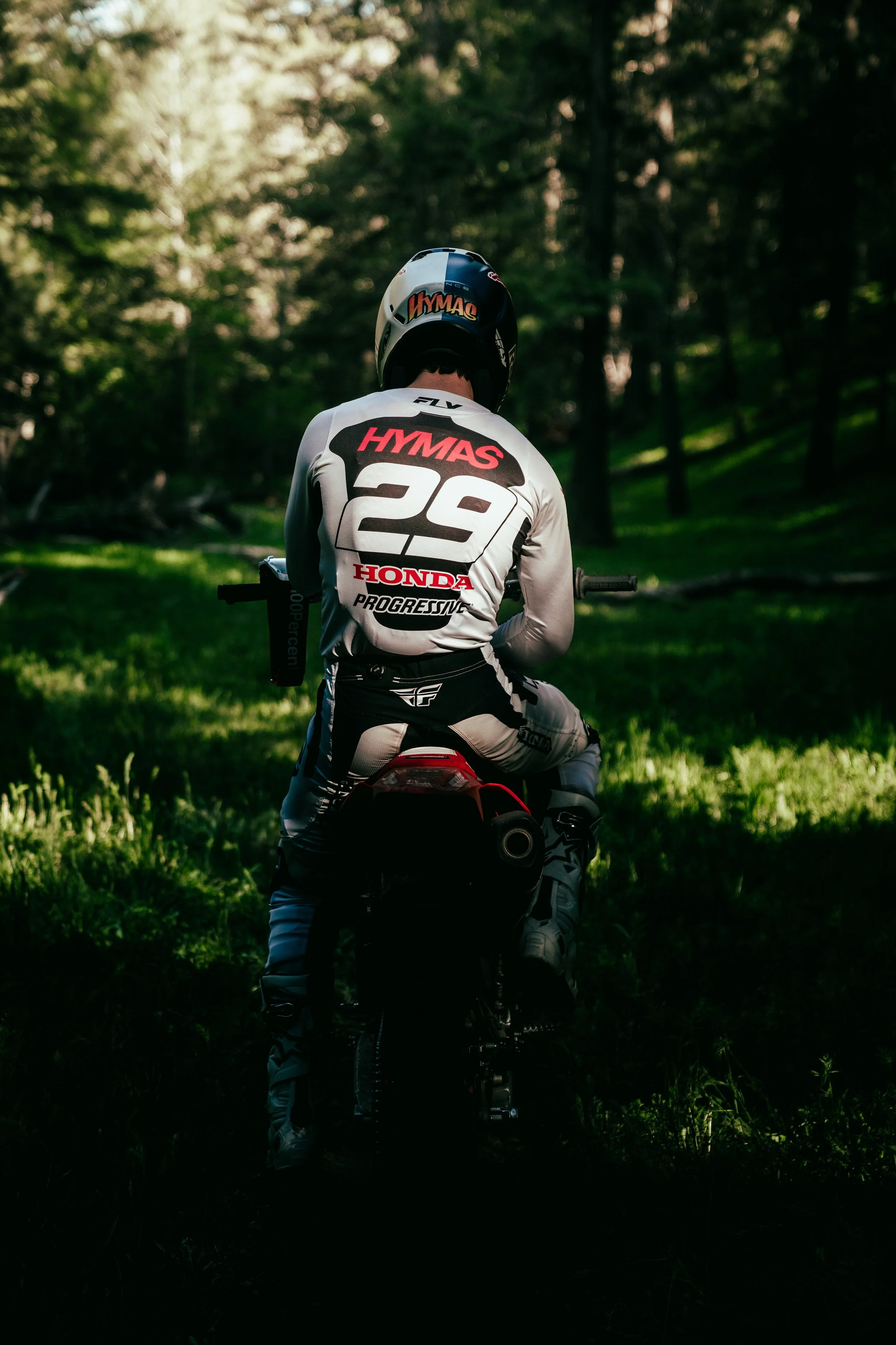 A person in racing gear and helmet riding a dirt bike through a forested area with sunlight filtering through the trees.