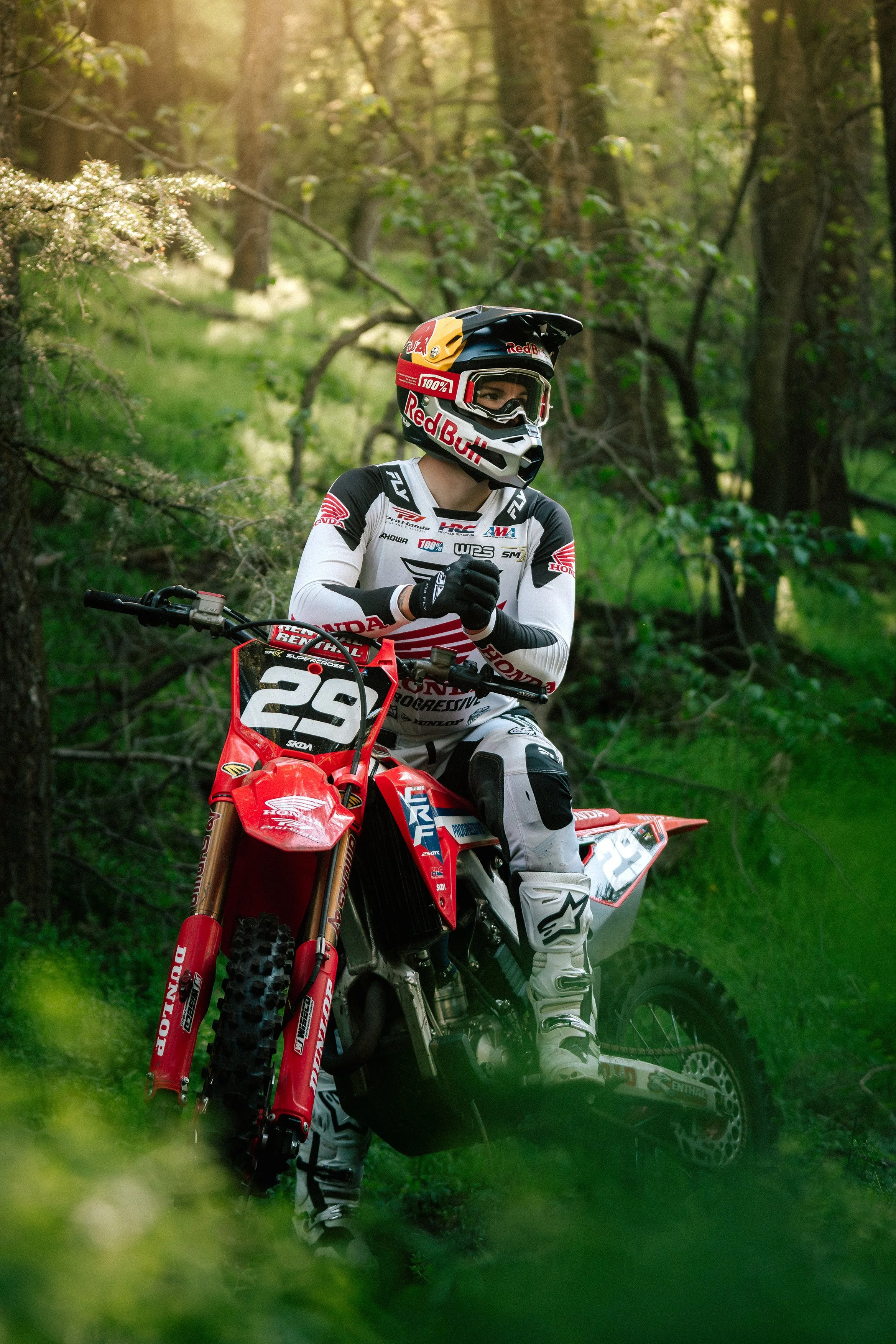 Motocross rider in a helmet and racing gear sitting on a red dirt bike in a forest, with sunlight filtering through trees.