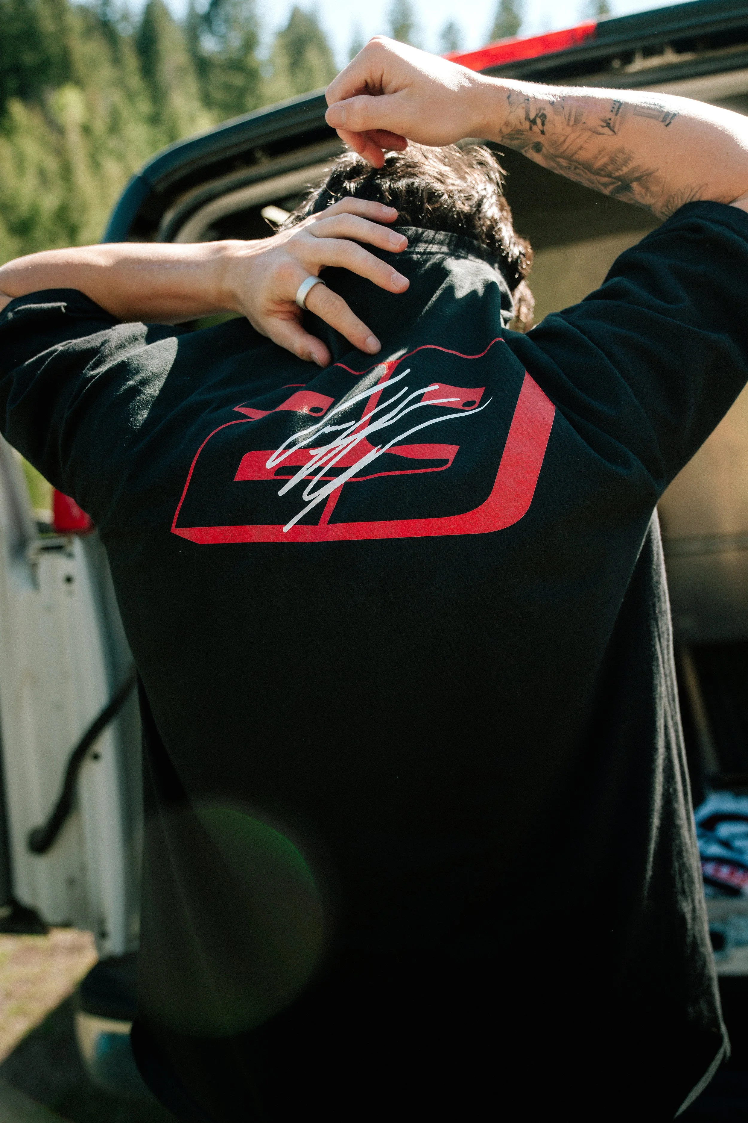 Person with tattoos adjusting hair in front of an open vehicle trunk in a wooded area.