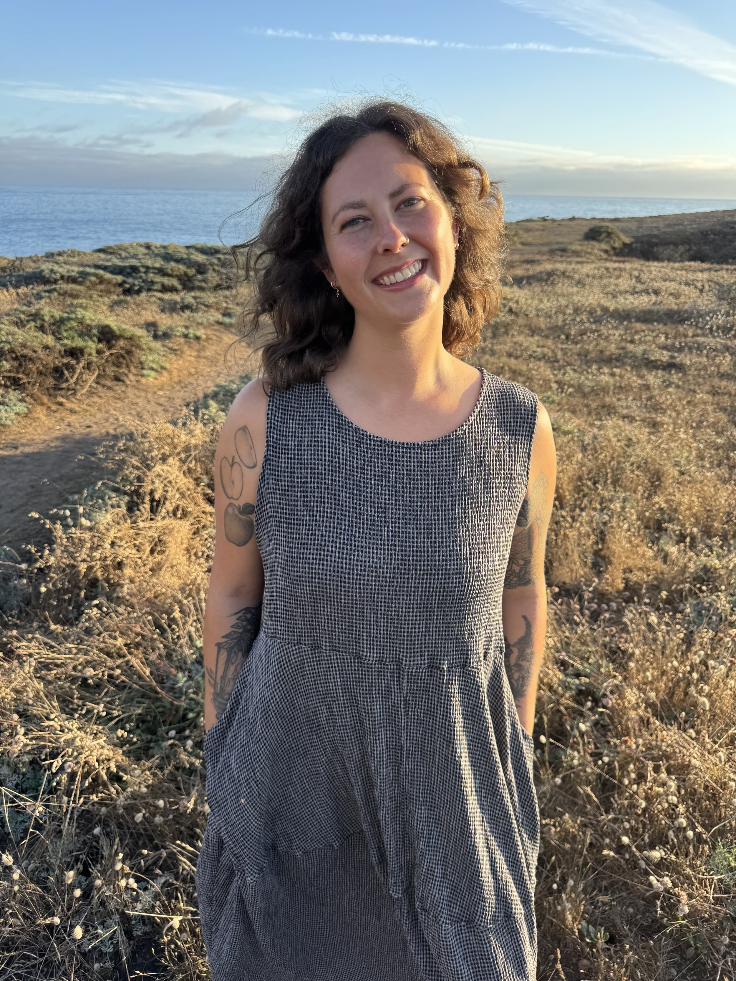 A smiling woman with curly brown hair standing outdoors near the coast during sunset, wearing a black and white checkered sleeveless dress with tattoos on her arms.