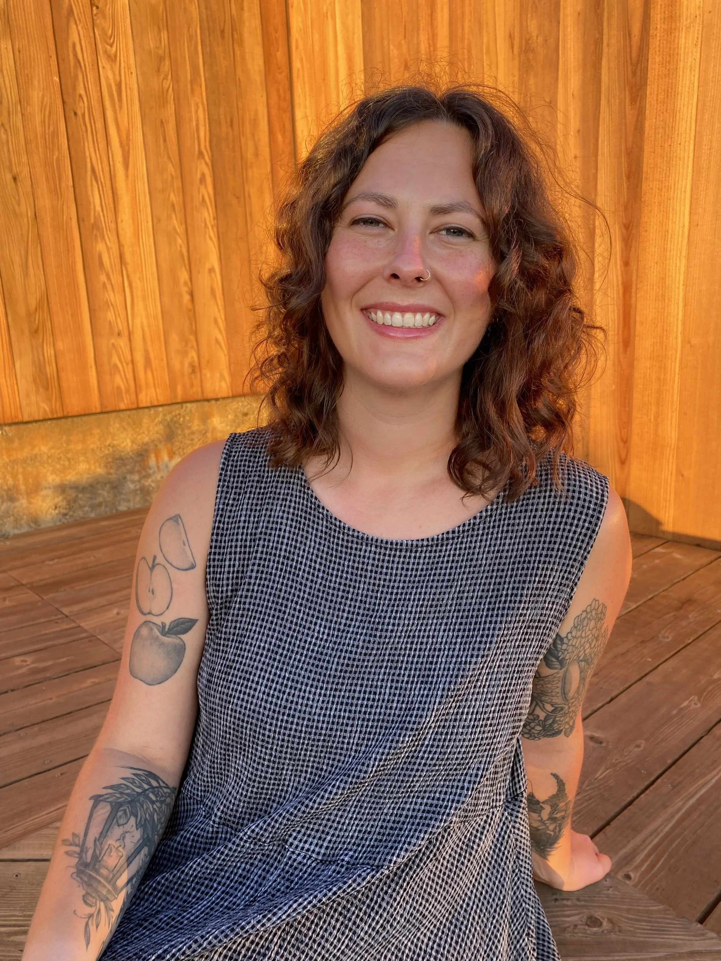 A woman with curly brown hair smiling, wearing a checkered sleeveless dress, seated outdoors on a wooden deck with a wooden fence in the background, displaying tattoos on her arms.