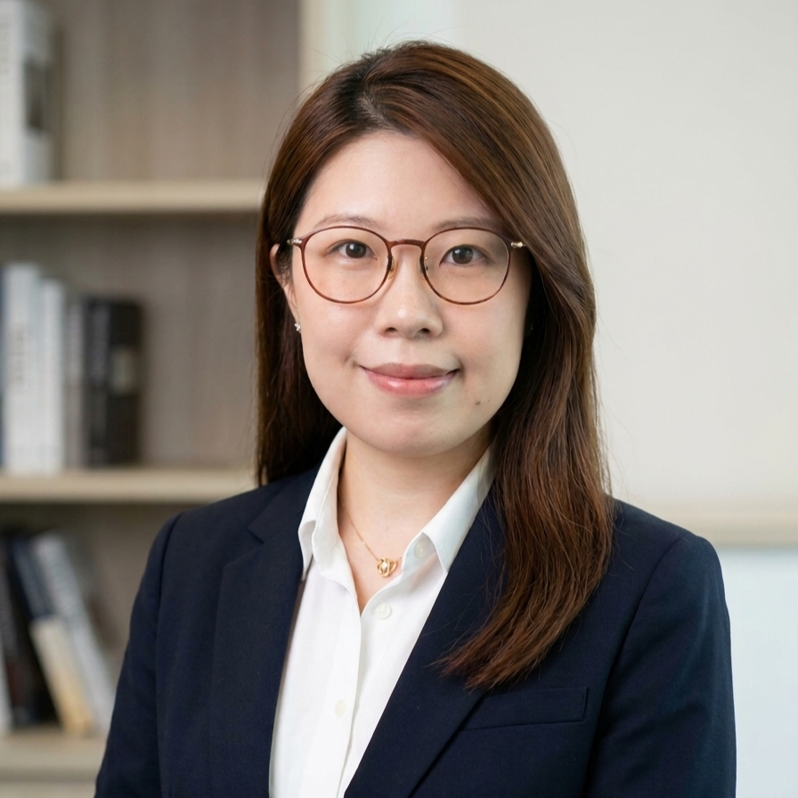 Professional woman with glasses and long brown hair, smiling in an office setting with bookshelves in the background.