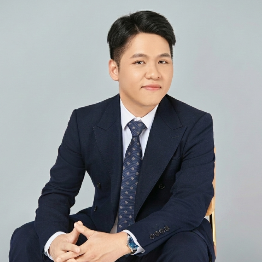 A young man in a navy blue business suit and tie sitting on a chair against a plain light gray background.