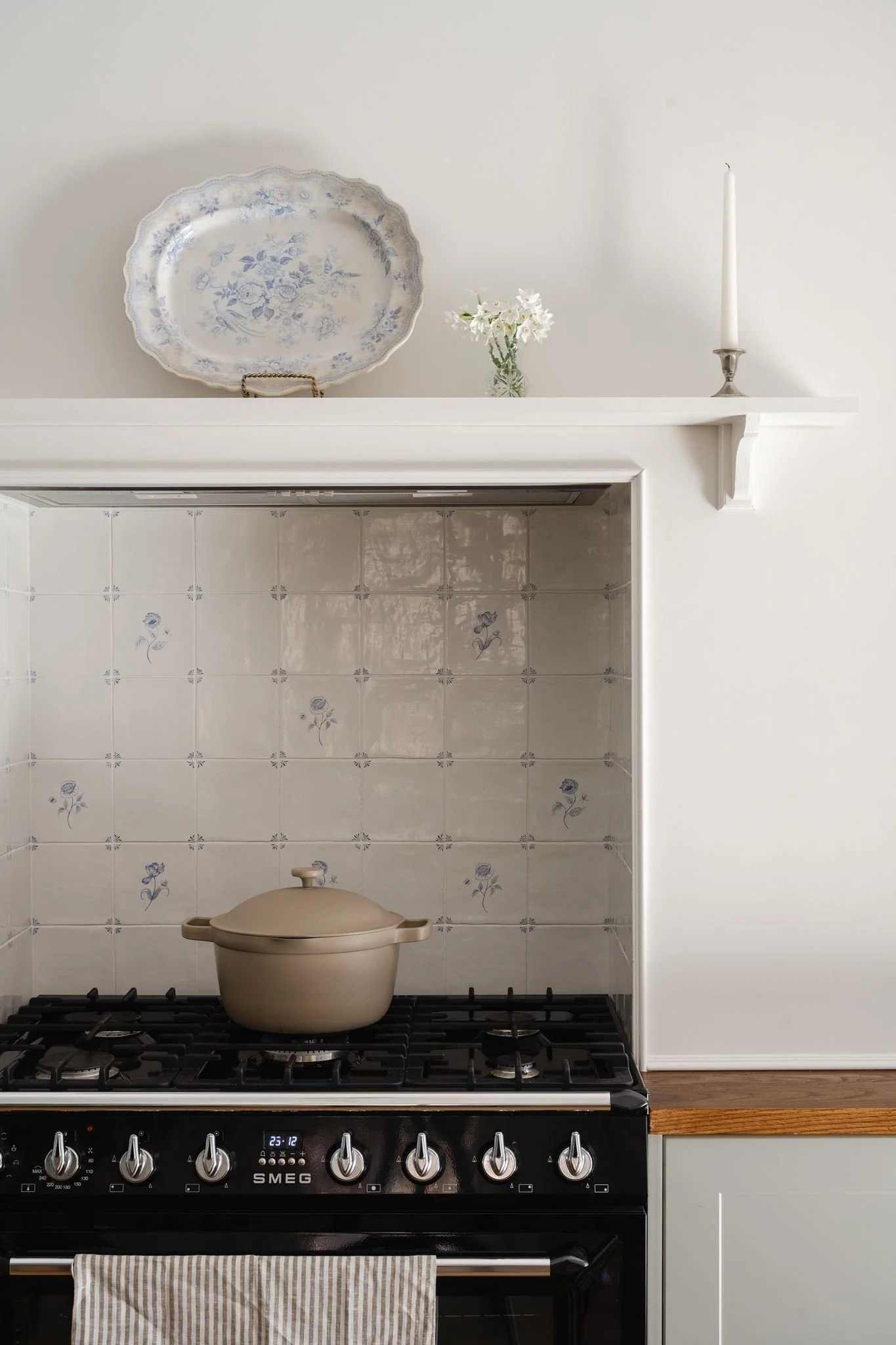 Kitchen scene with a black SMEG gas stove, a beige pot on one burner, beige cabinets, and a white countertop. A striped towel hangs from the oven handle. The upper shelf holds a decorative porcelain platter with blue floral designs, a small glass vas