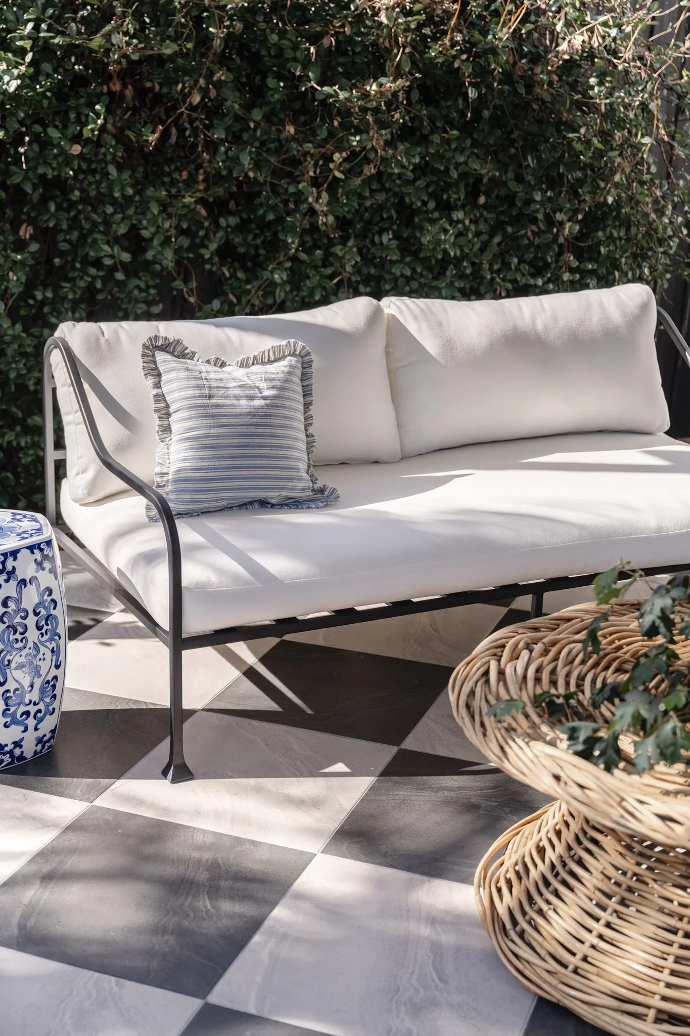 A white outdoor sofa with a striped pillow, situated on a checkered black and white tile patio, with a lush green hedge in the background and a decorative blue and white ceramic side table, along with a woven wicker basket with greenery.