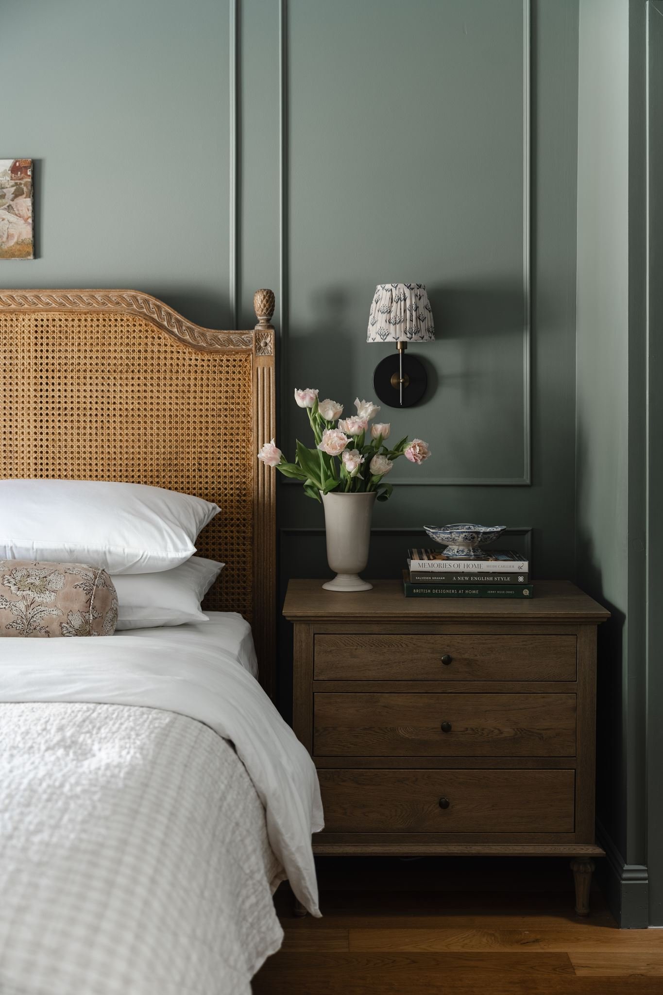 A bedroom corner with a wooden bed with a woven headboard, a wooden nightstand with a stack of books and a ceramic bowl, a lamp with a patterned shade, a vase with pink flowers, and a wall with green paneling.