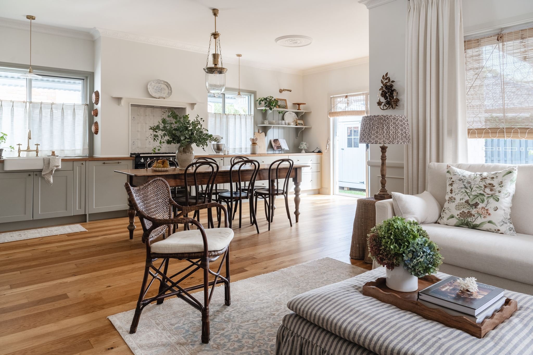 A bright and cozy open-plan living and kitchen area with wooden floors, white walls, and large windows with curtains. The space features a dining table with six chairs, a white sofa with floral and striped cushions, a wooden side table with a potted 