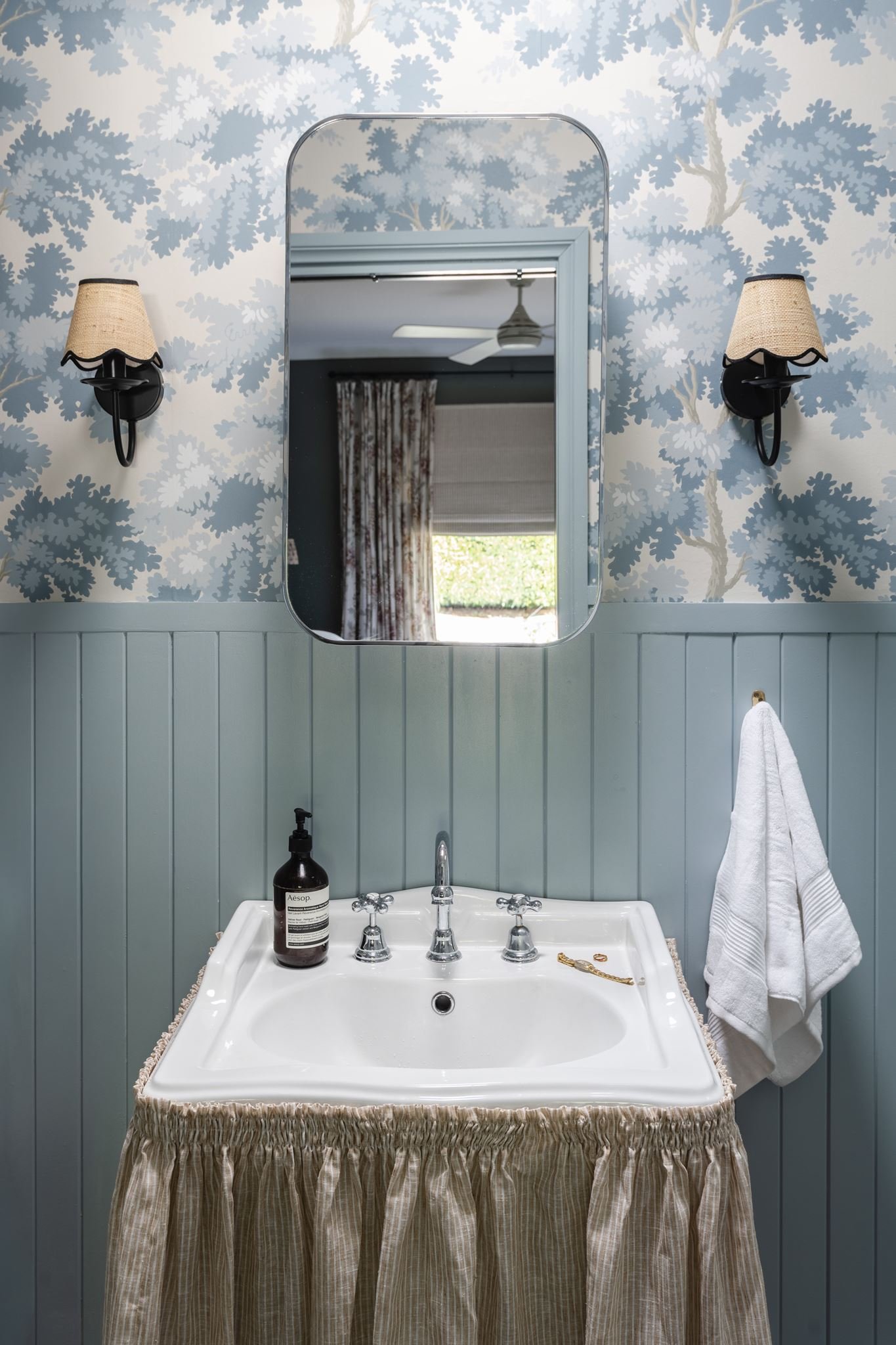 Bathroom sink with a beige skirted curtain, a white soap dispenser, a mirror, and wall sconces with beige shades. Towel hanging on wall hook and ceiling fan visible in reflection.