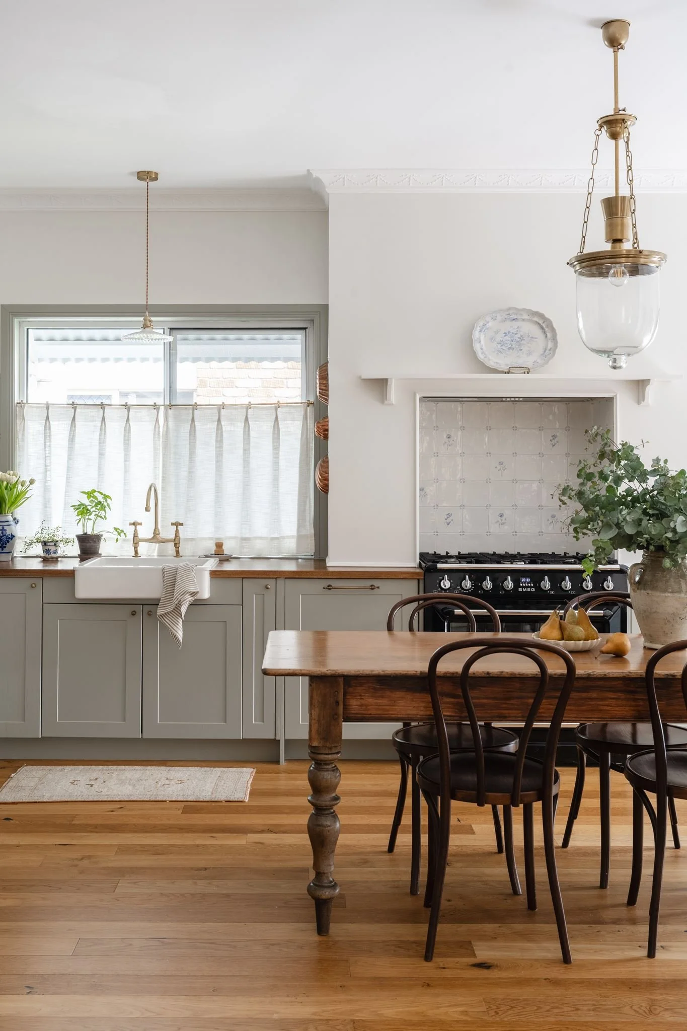 A cozy kitchen with light gray cabinets, a white farmhouse sink, green plants on the windowsill, and a wooden dining table with black chairs. There are two pendant lights hanging from the ceiling, and a decorative plate on a white shelf above the stove area. The kitchen has a warm wood floor and a natural, inviting atmosphere.
