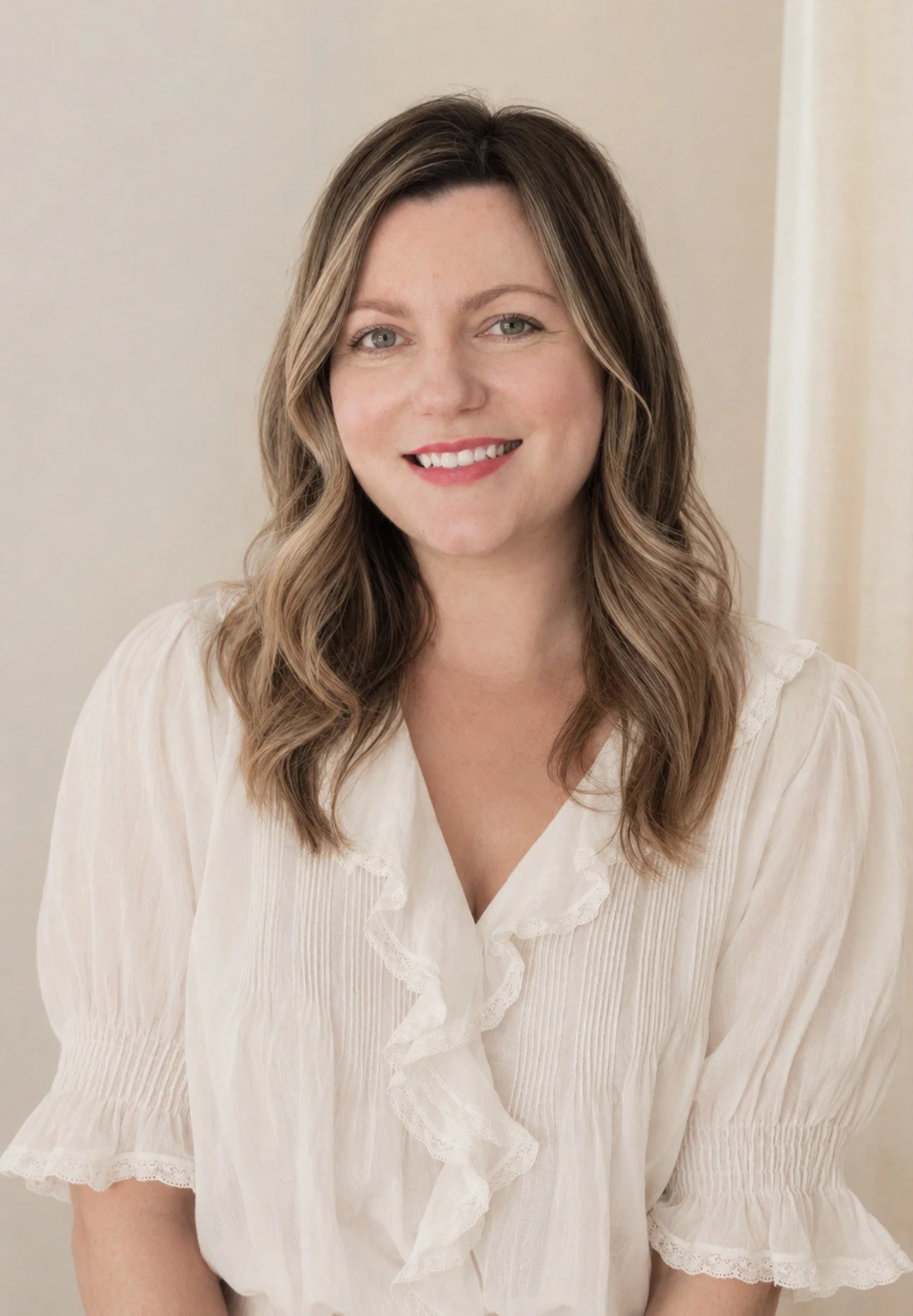 A woman with wavy brown hair and fair skin smiling, wearing a light-colored blouse with ruffled collar and sleeves, in front of a beige background.