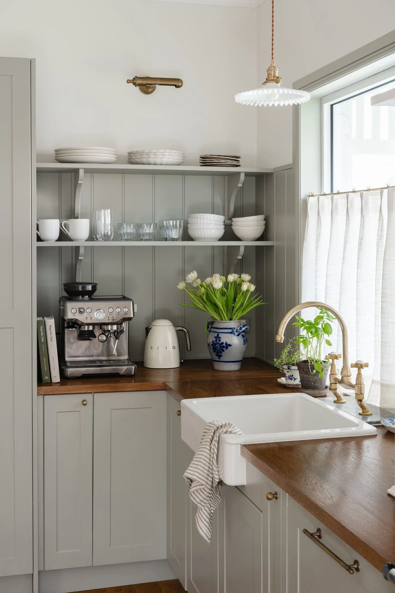 Kitchen with open shelves holding white dishes and glasses, a coffee machine, electric kettle, plants on the counter, a kitchen sink, and a window with curtains.