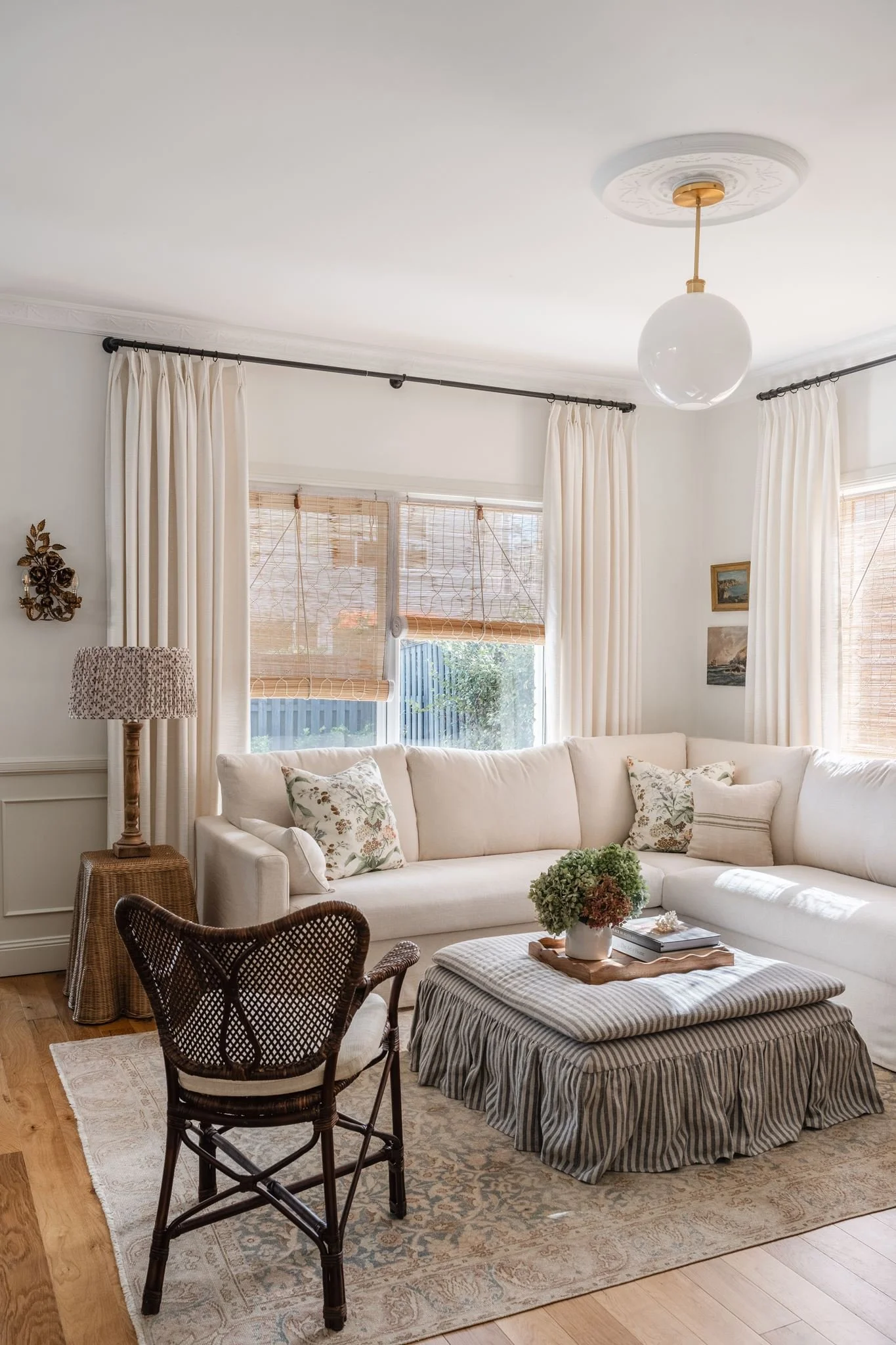 A cozy living room with a white sectional sofa decorated with floral pillows, a striped ottoman with a flower arrangement, a rattan chair, and a wooden side table with a lamp. The room features cream curtains, bamboo blinds, ceiling molding, and a mo