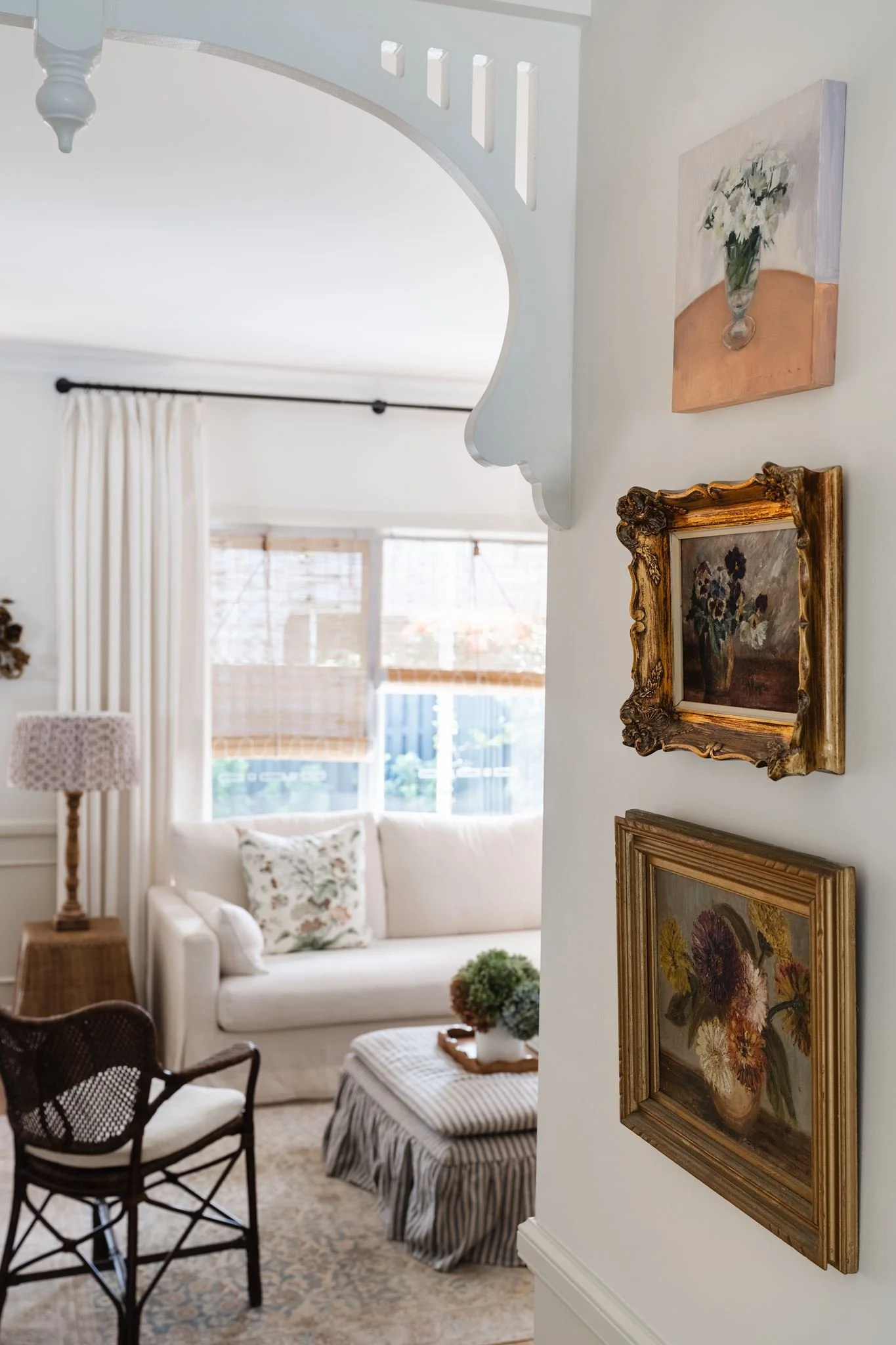Living room with white sofa, floral pillows, striped ottoman, wooden side table with flowers, and art on the wall, cream-colored walls, large window with white curtains, and patterned rug.